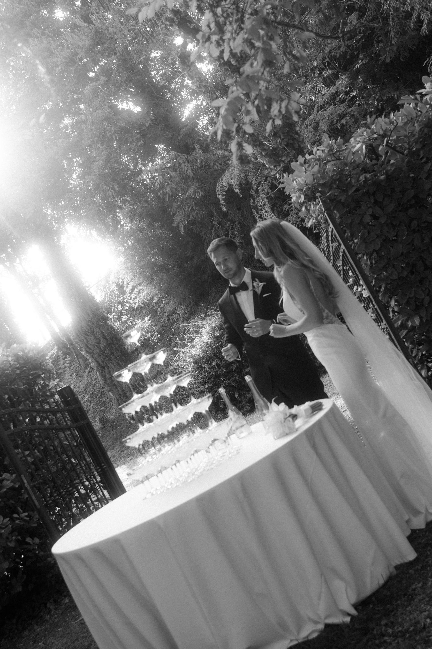 Black and white photo of a couple pouring champagne into stacked glasses outdoors.