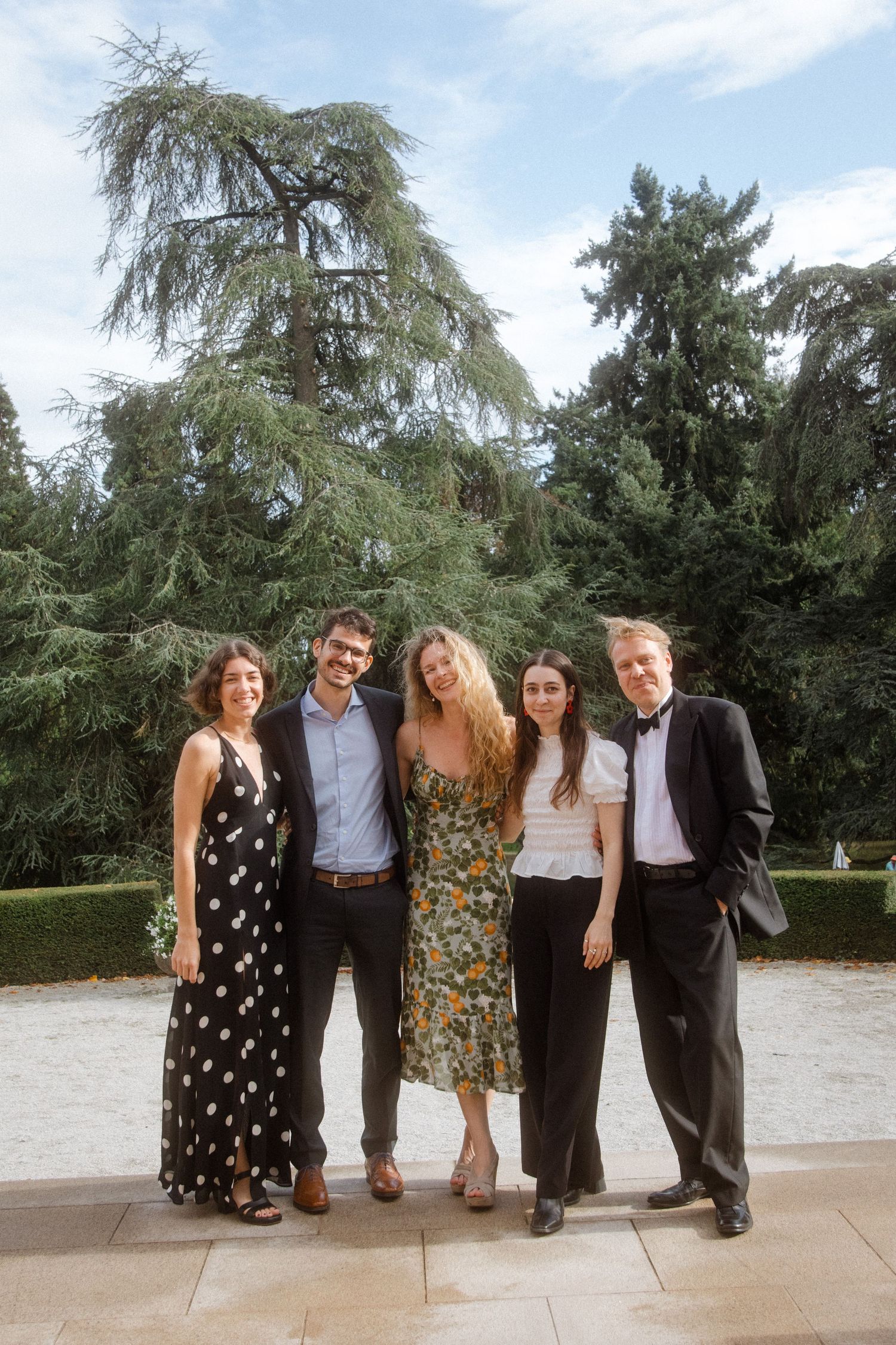 A group in formal attire poses together outdoors against a backdrop of trees.