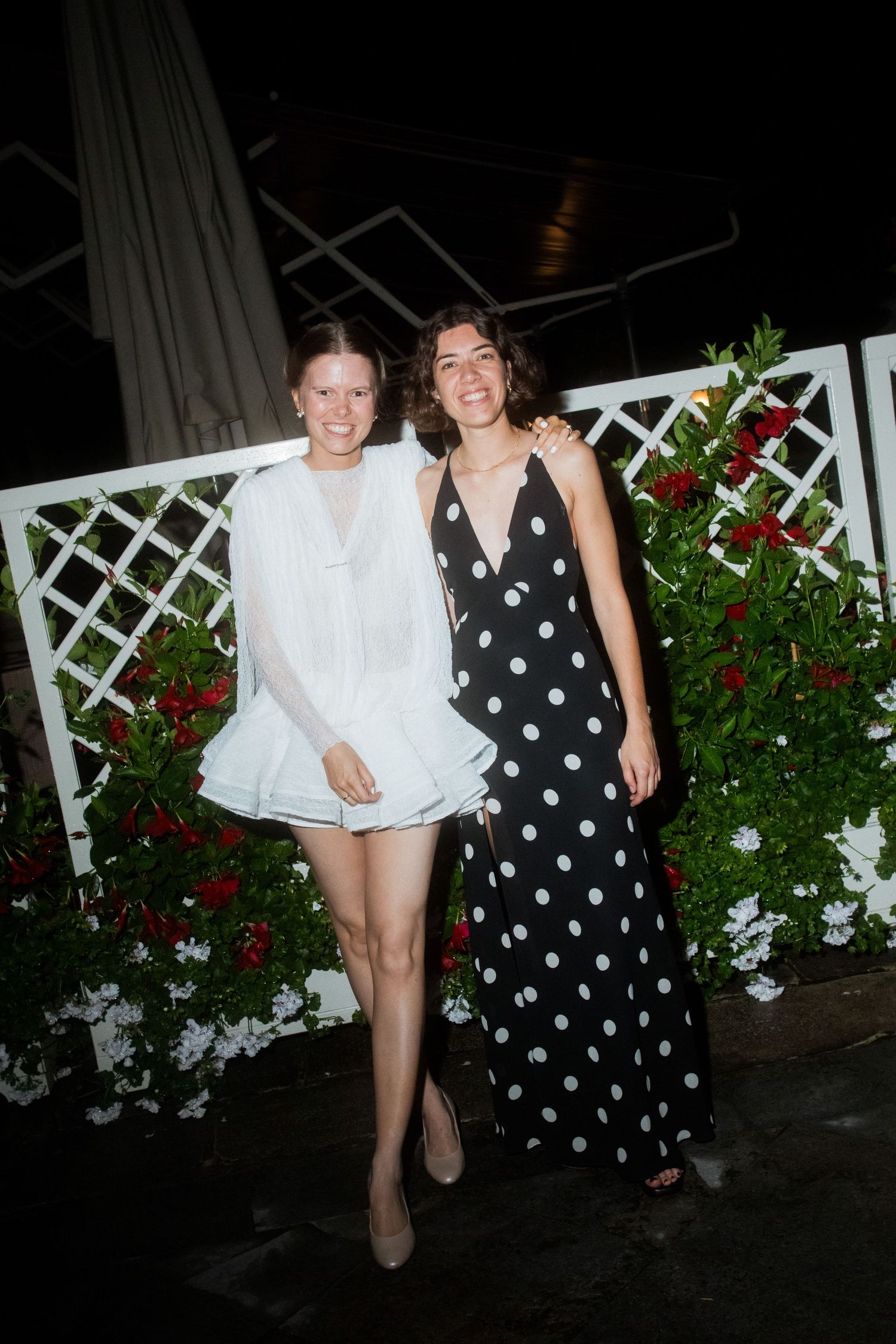 Two people posing together outdoors by white lattice and red flowers at night.