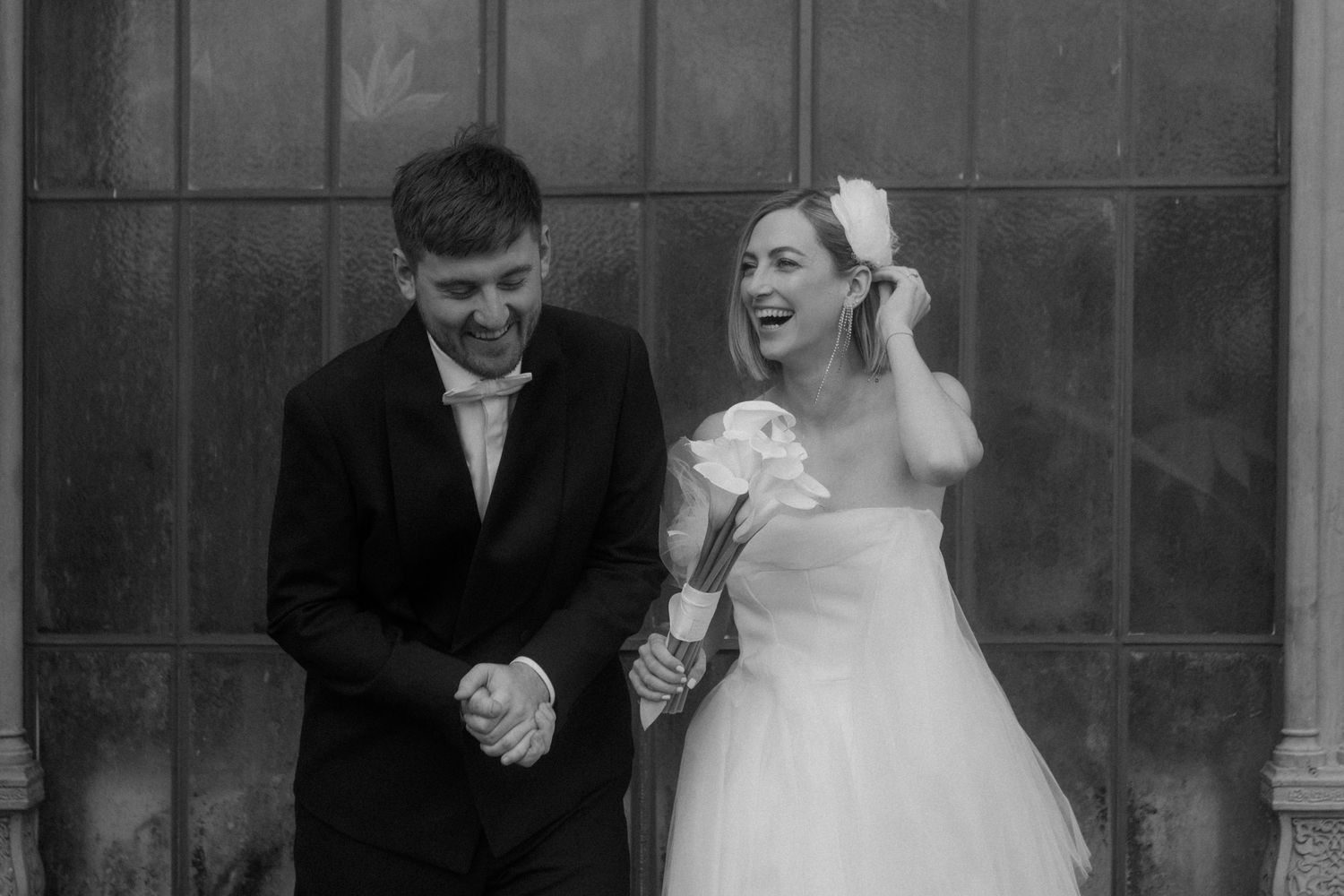 Black and white candid shot of joyful newlyweds laughing together against stone wall.