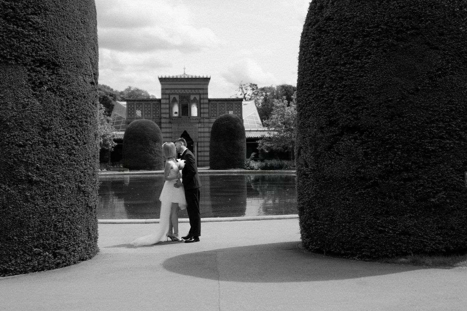 Black and white photo of a couple embracing between manicured hedges near a reflecting pool.