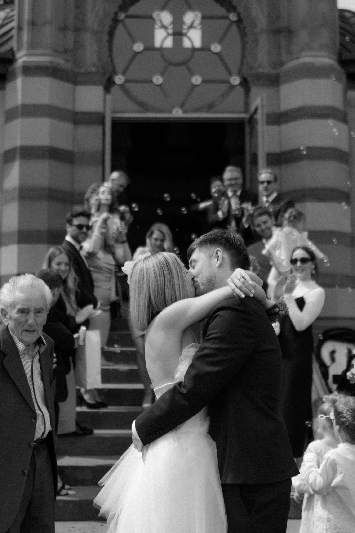 Black and white photo of a wedding couple embracing on church steps while guests celebrate.