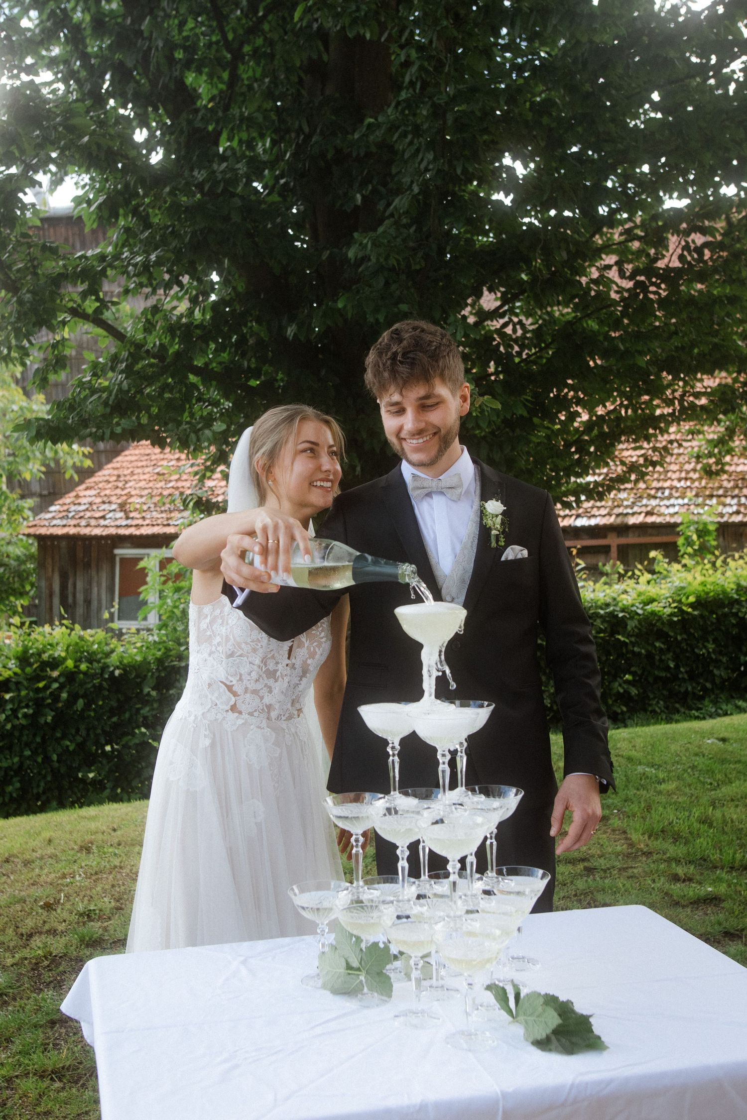 Wedding couple pouring champagne into elegant crystal glass tower outdoors.