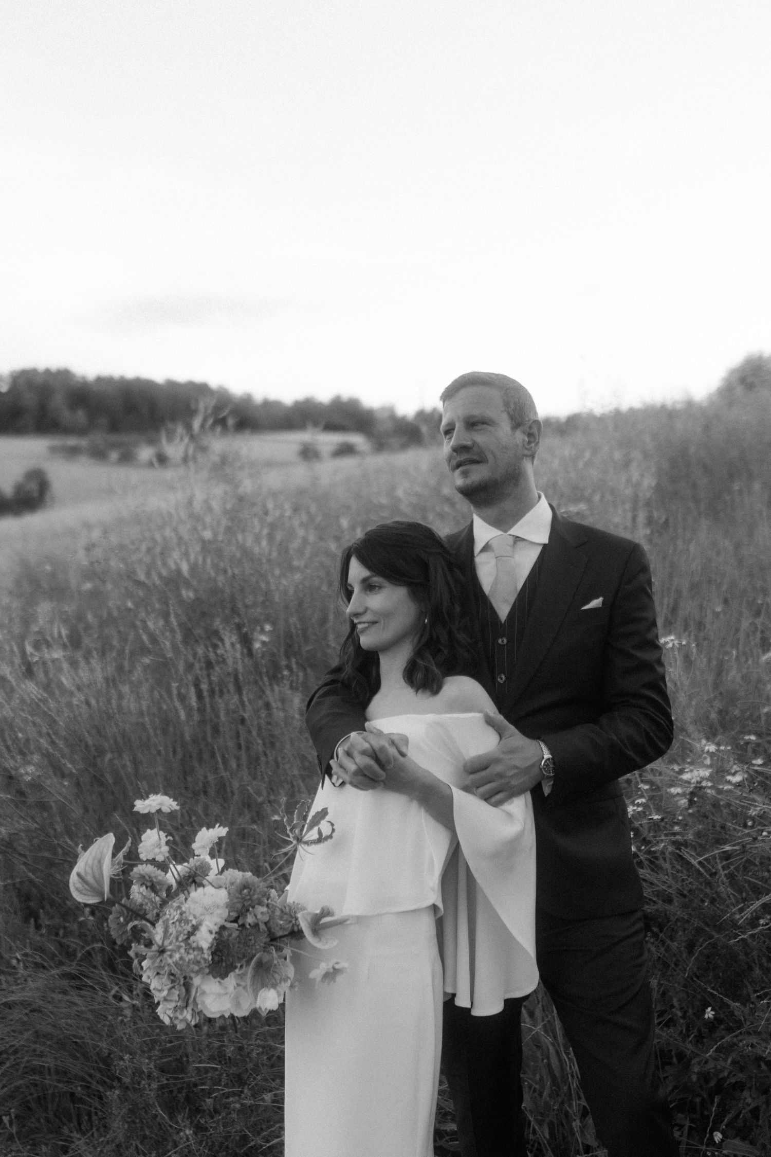 A black and white portrait of newlyweds embracing in a summer field.