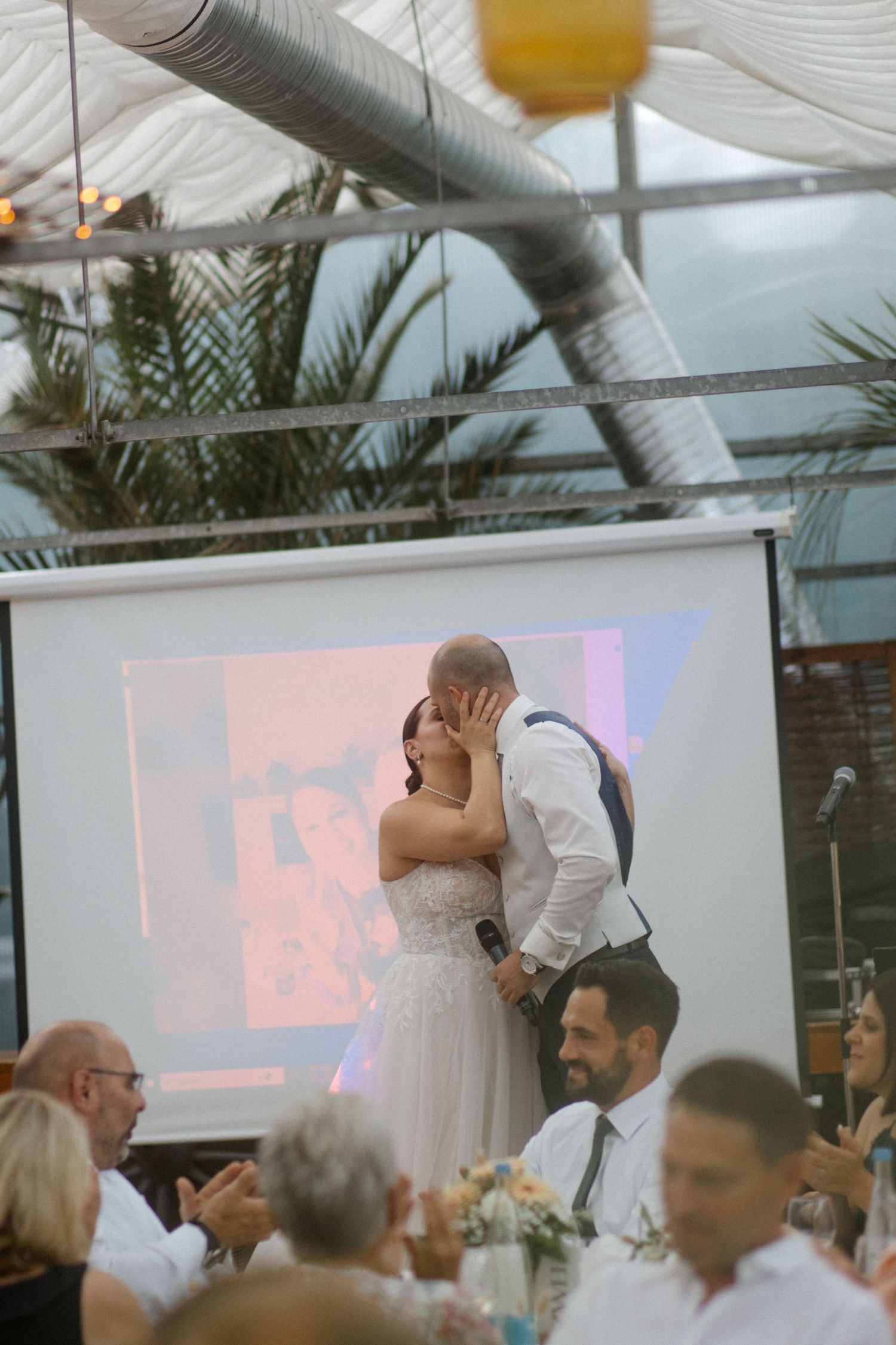 A tender wedding kiss captured in an indoor reception venue with palm trees and string lights overhead.