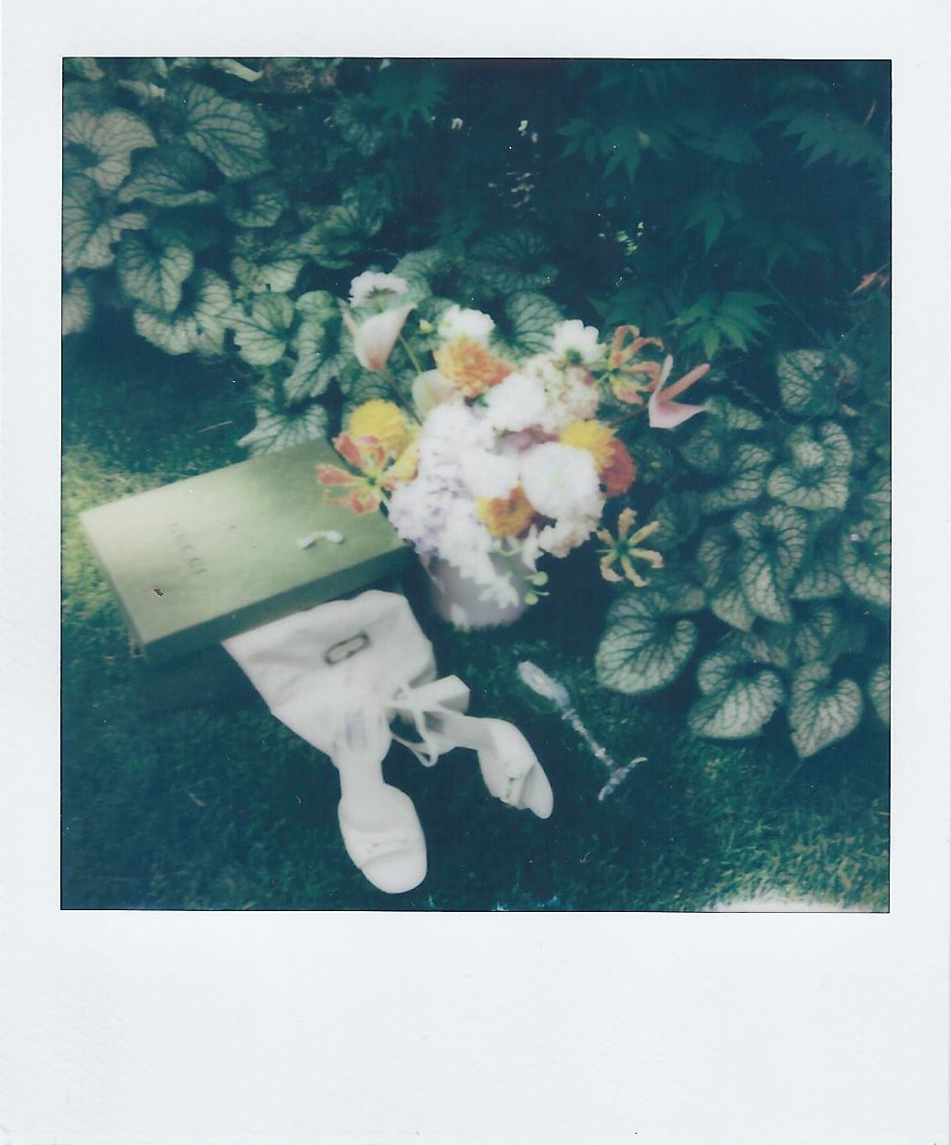 A bridal bouquet, white shoes and other wedding items arranged on grass with green foliage.