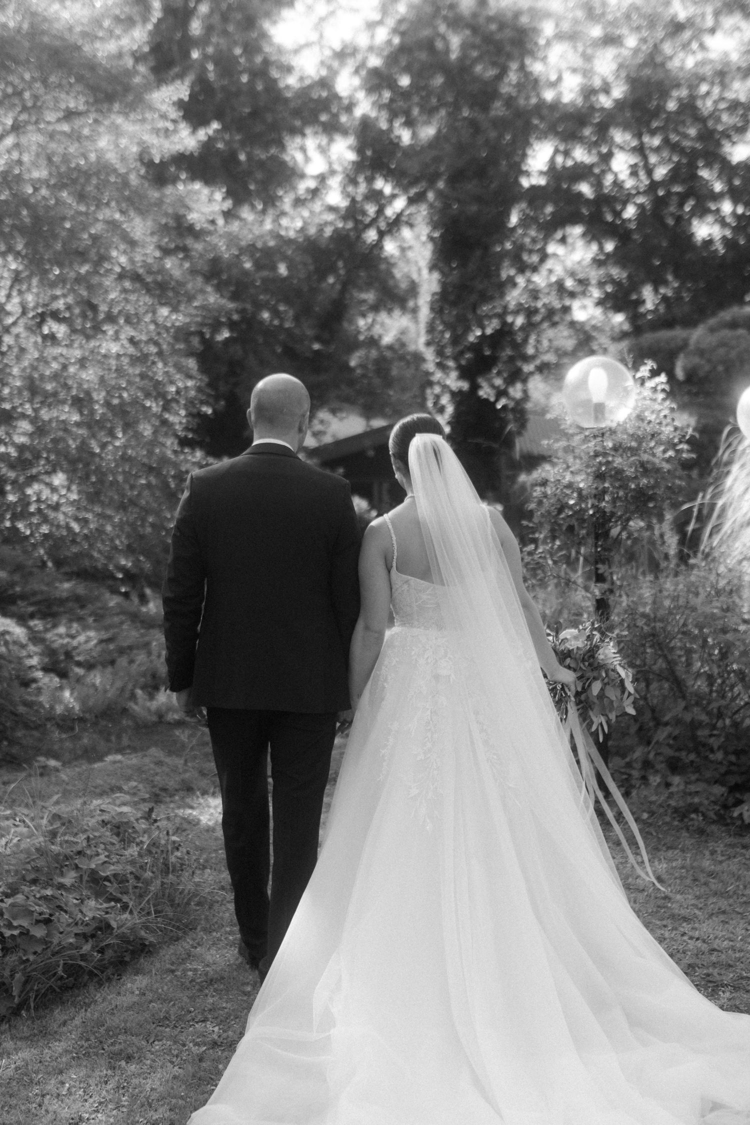 Black and white photo of newlyweds walking along a garden path surrounded by lush greenery.