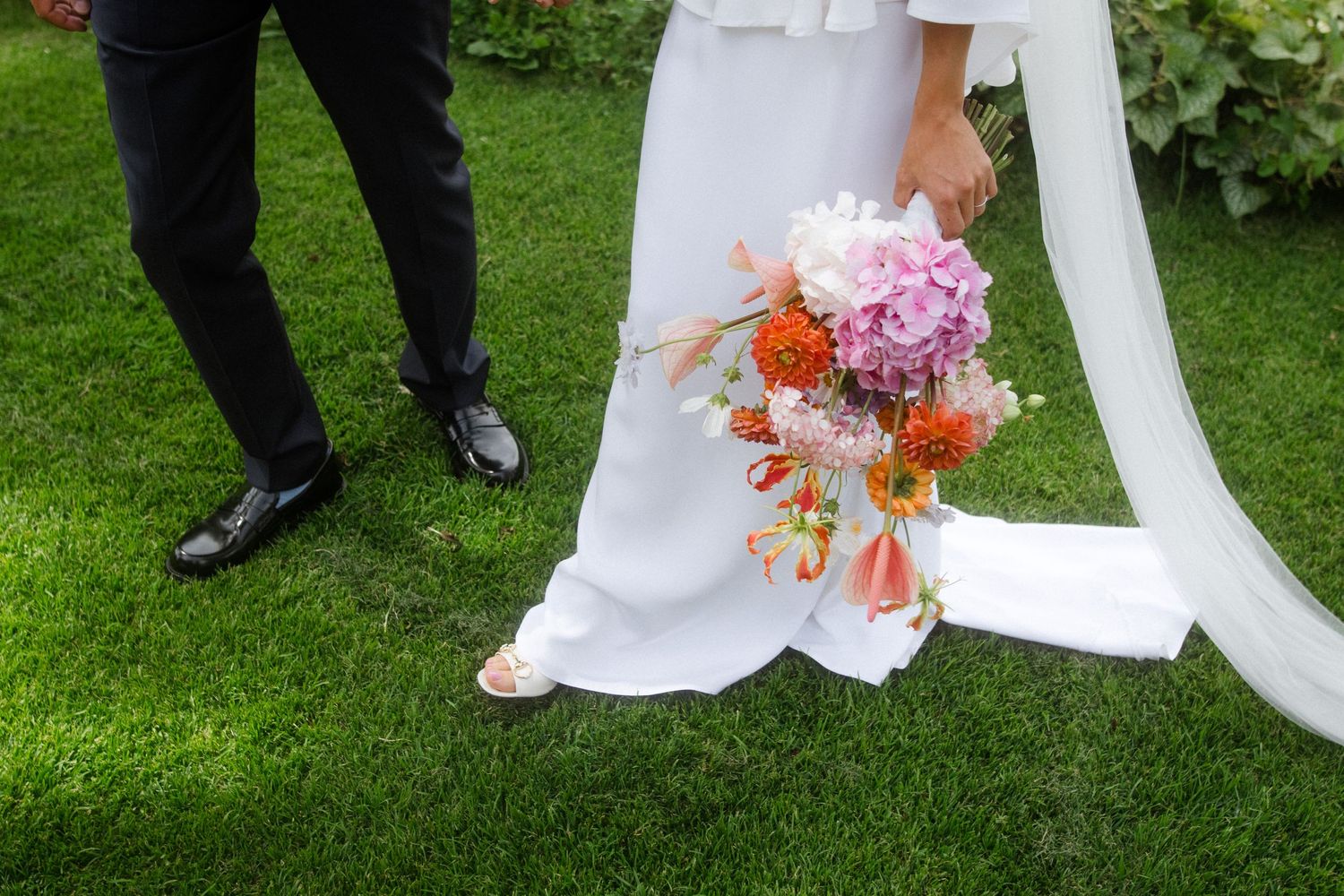 Wedding dress hem and formal shoes on grass with vibrant pink and orange floral bouquet.