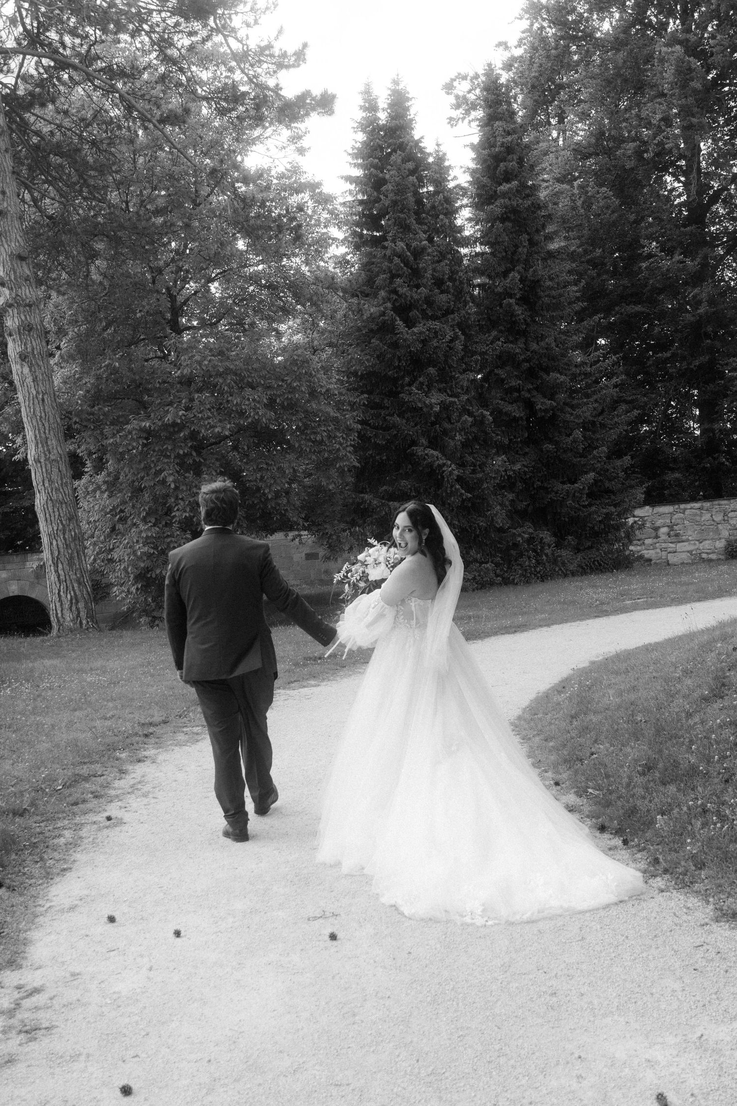 A couple walking along a tree-lined path in formal wedding attire with a flowing white dress trailing behind.