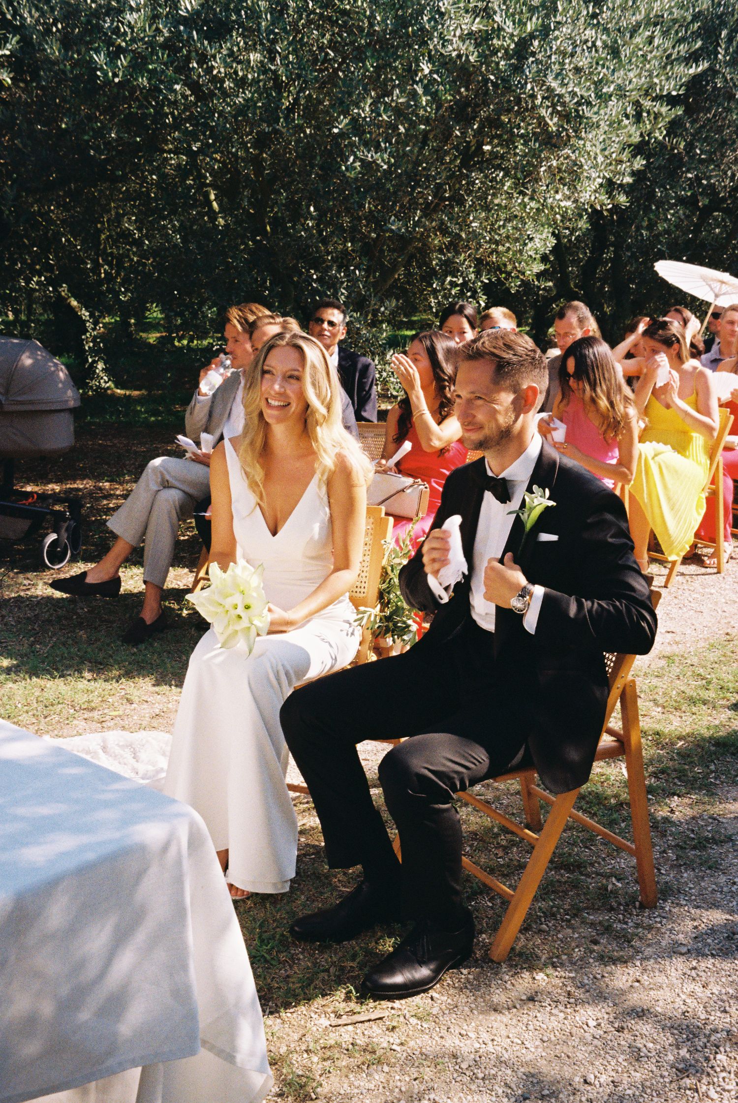 Wedding ceremony guests seated on wooden chairs in a garden setting.