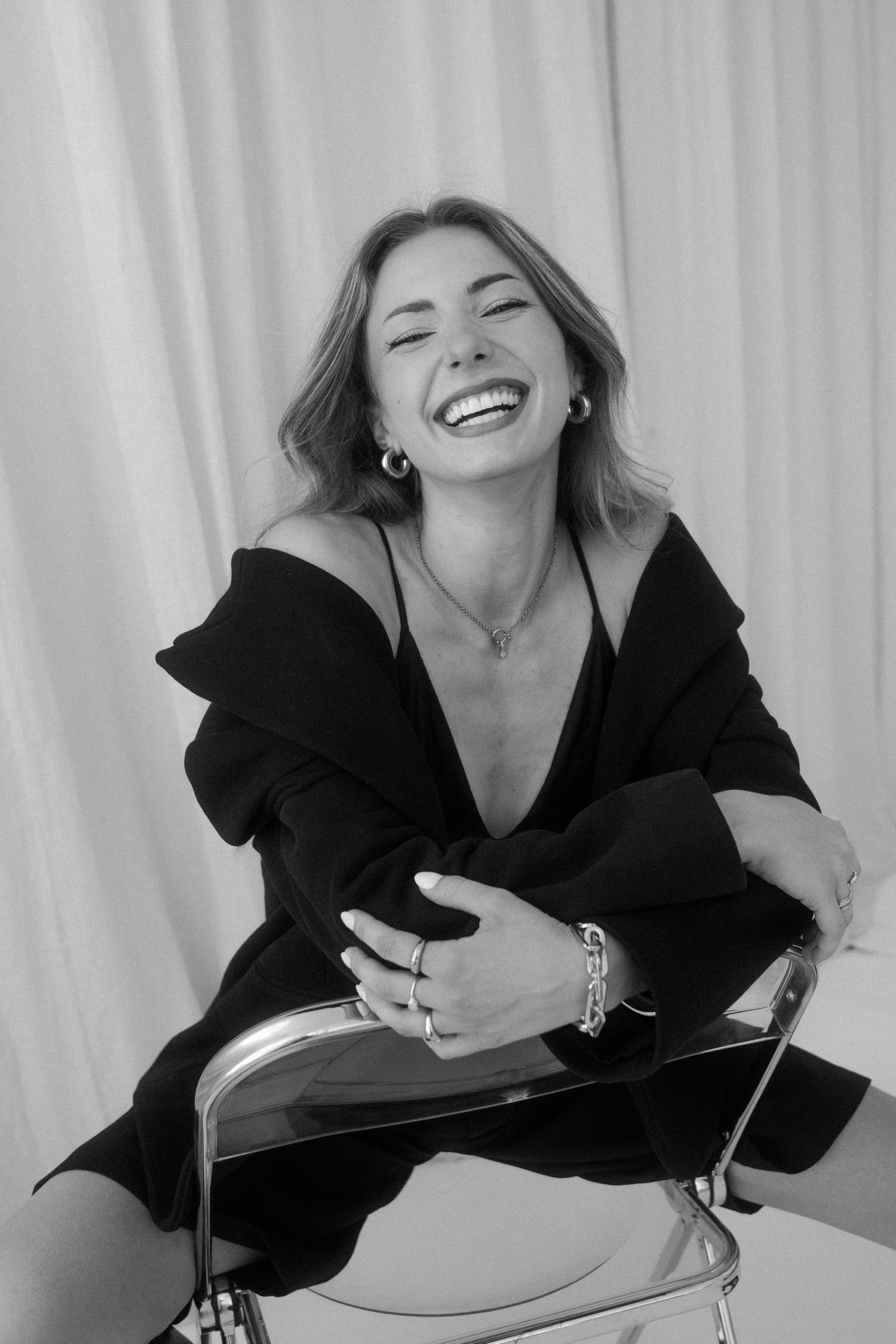 Black and white photo of a laughing woman in a black off-shoulder outfit, sitting on a chair wearing silver jewelry.