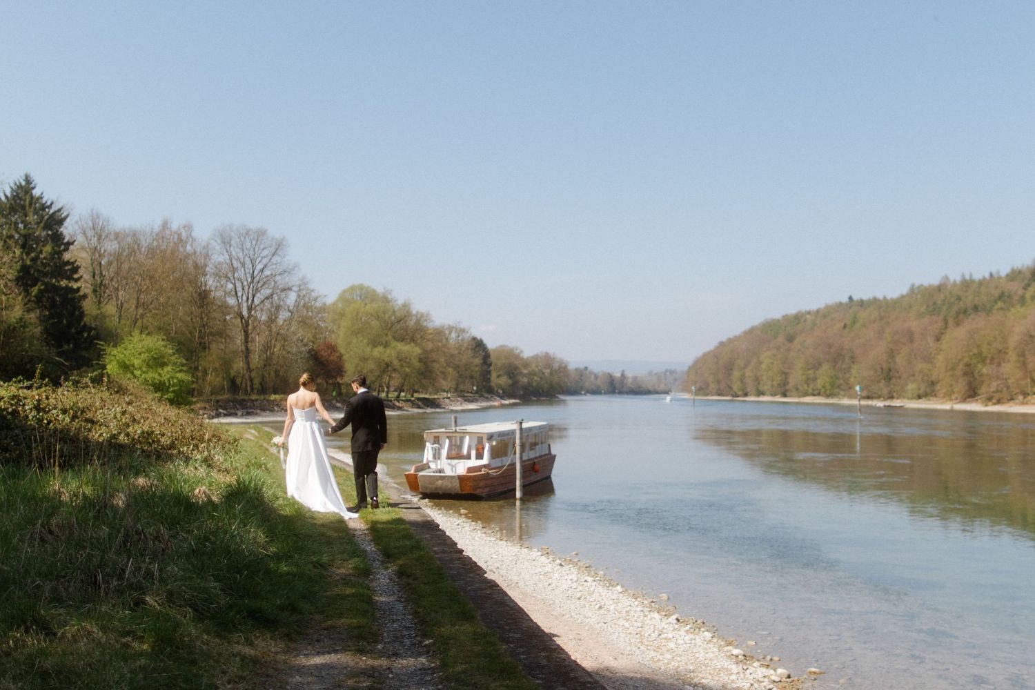 Bride and groom standing by a calm river with a boat docked nearby, surrounded by lush green trees on a clear day.