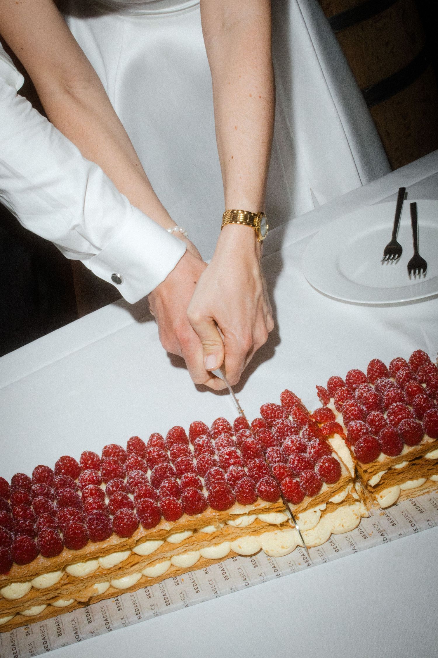 Close-up of bride and groom hands cutting a long rectangular raspberry mille-feuille wedding cake with a knife.