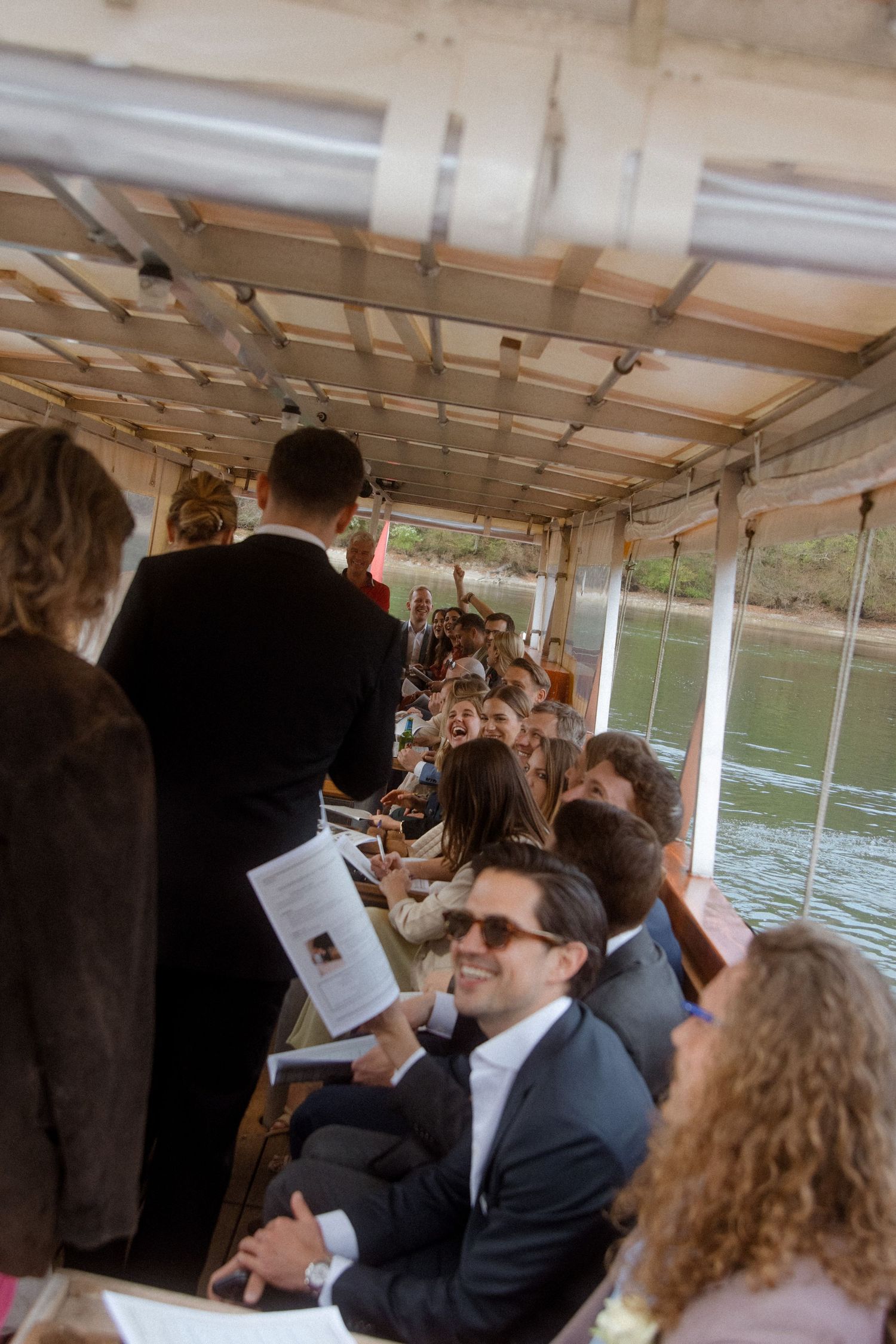 Wedding guests seated on a boat during a ceremony, smiling and holding programs, with water visible through windows.