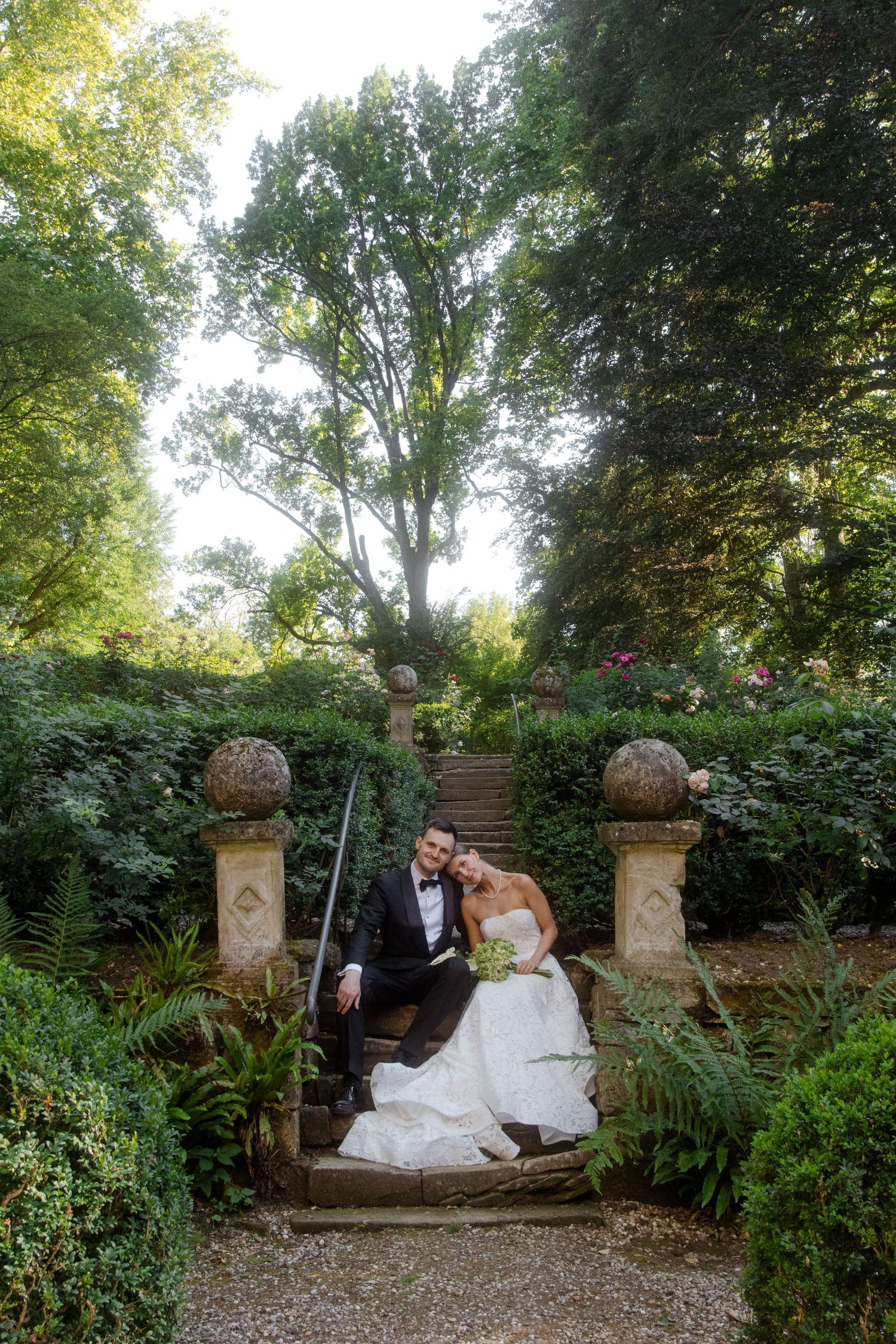 Bride and groom pose on stone garden steps surrounded by lush greenery in an elegant outdoor wedding portrait.