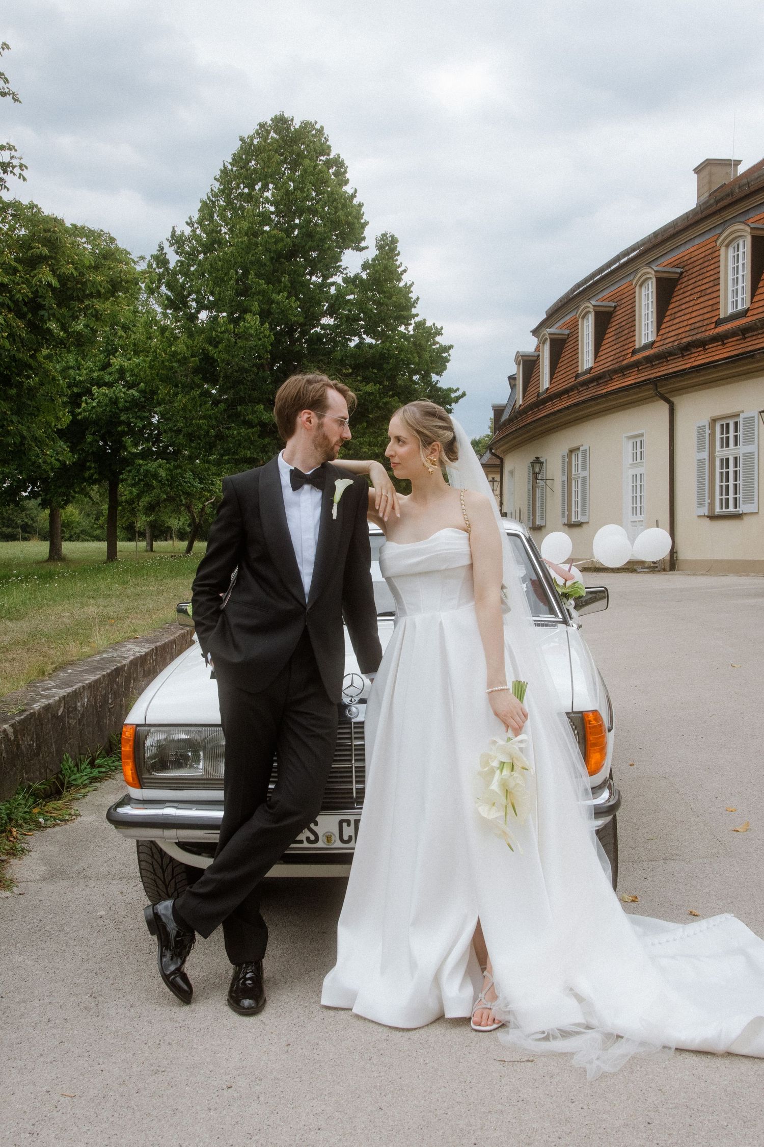 Newlyweds pose together near a classic car outside a historic building.