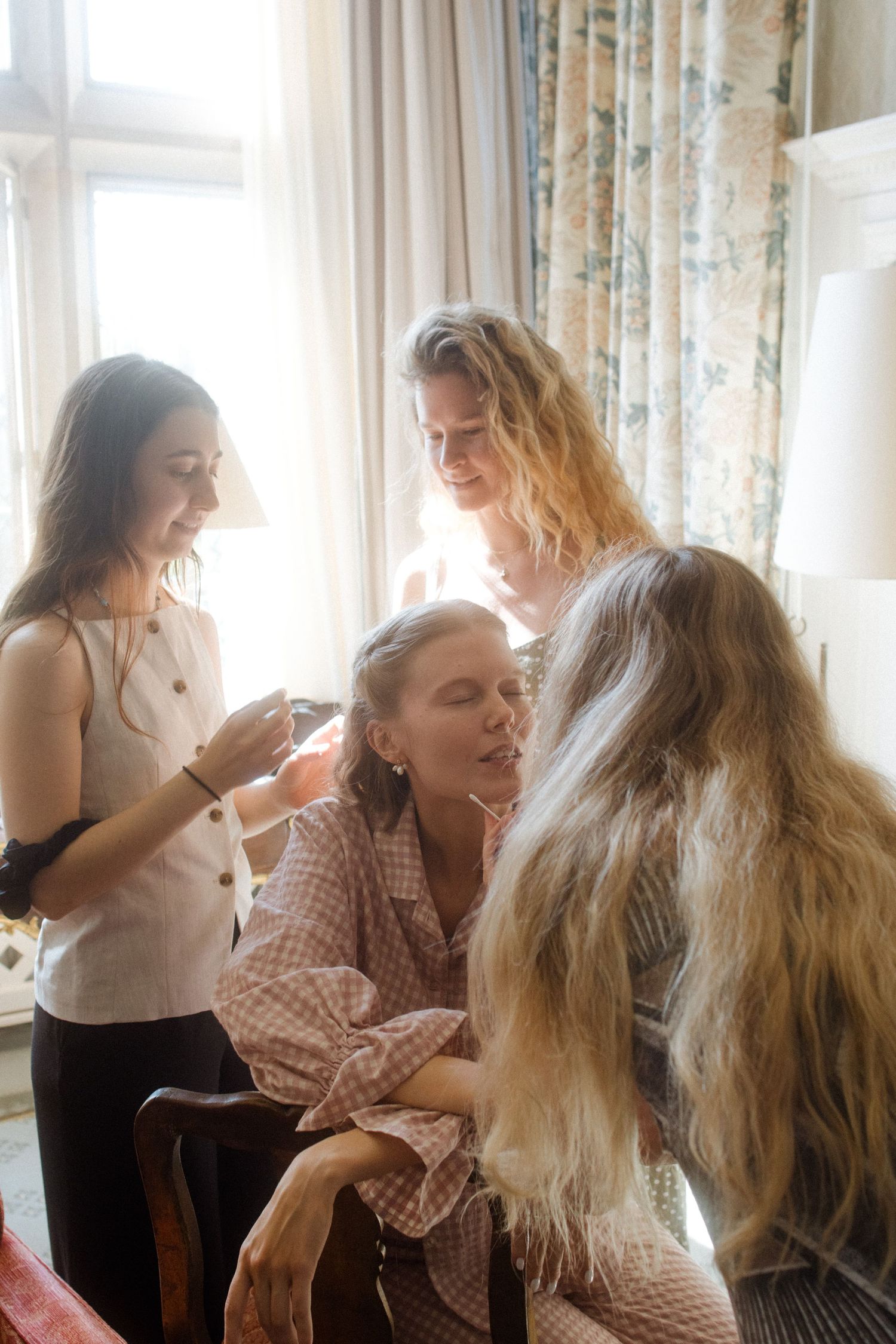 Two women with long hair playfully interact in a bright sunlit room with floral curtains.
