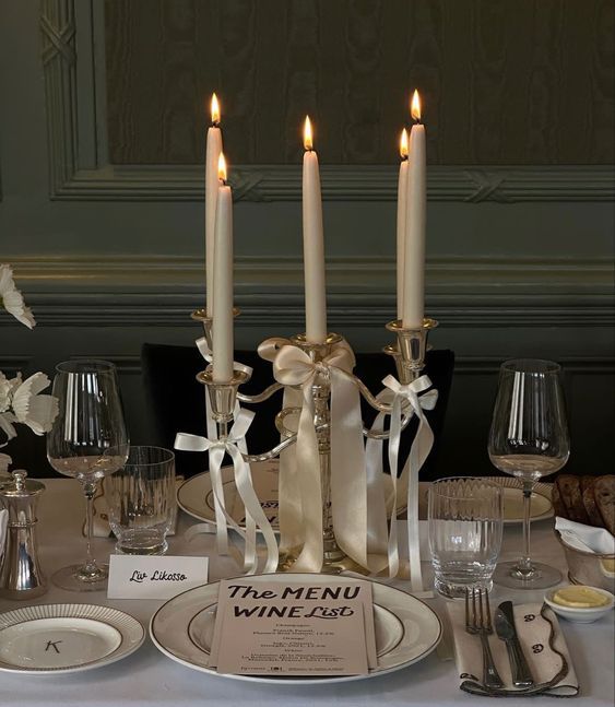Elegant table setting with white candles, ribbon bows, and wine glasses against a dark green wall.
