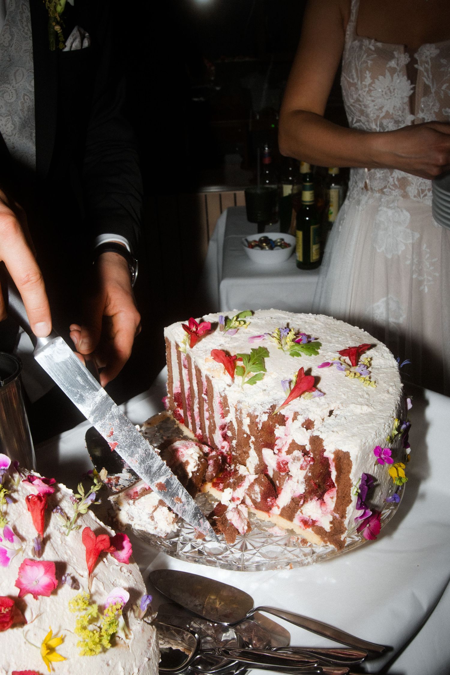 A white wedding cake decorated with flowers and strawberries being cut during a reception celebration.