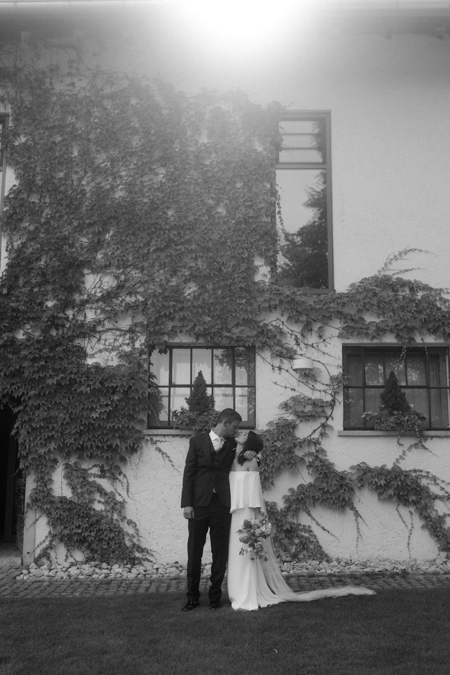 Black and white portrait of newlyweds standing by vine-covered building.