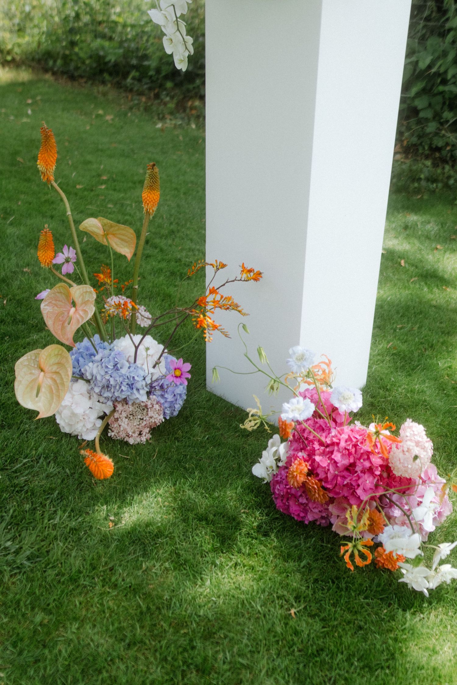 Elegant floral arrangements with hydrangeas and orange accents decorating white pillars on grass.