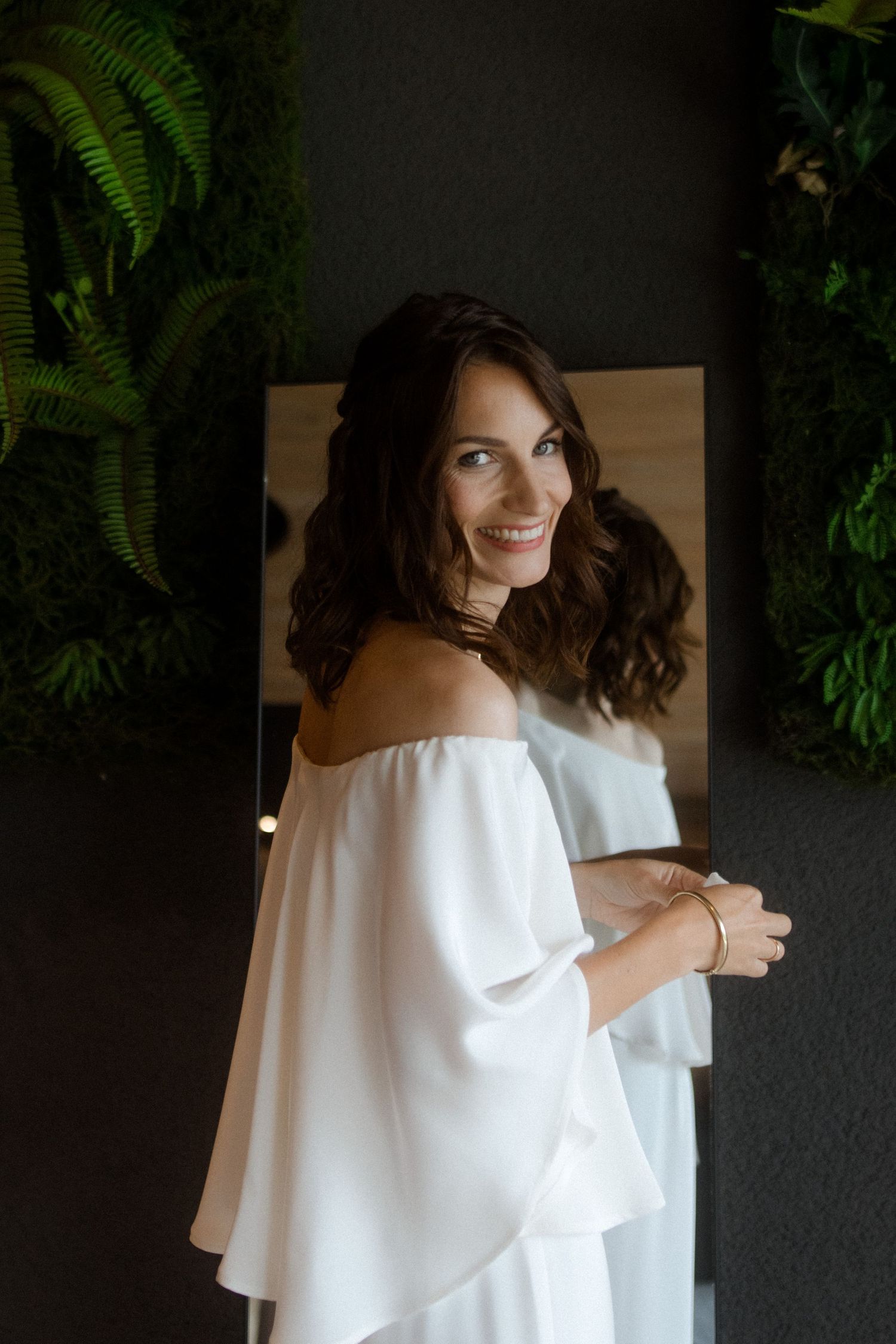Woman in white off-shoulder dress stands against dark wall with ferns.
