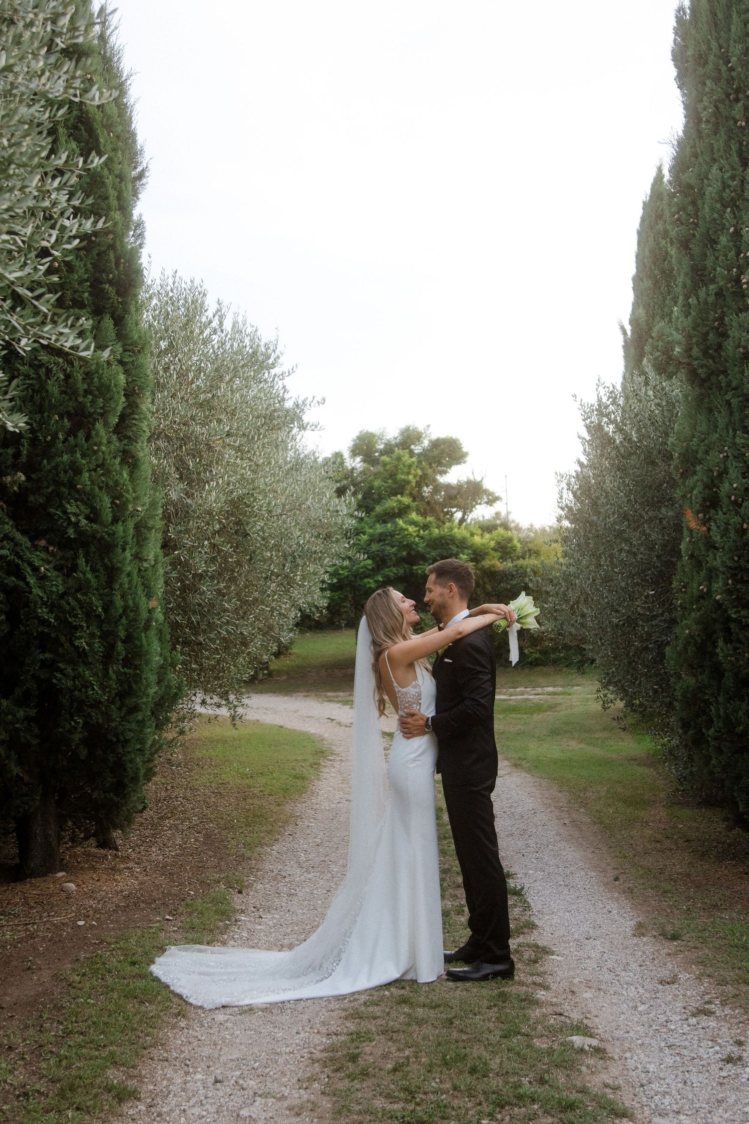 A romantic wedding couple embraces on a cypress-lined path in an elegant garden setting.