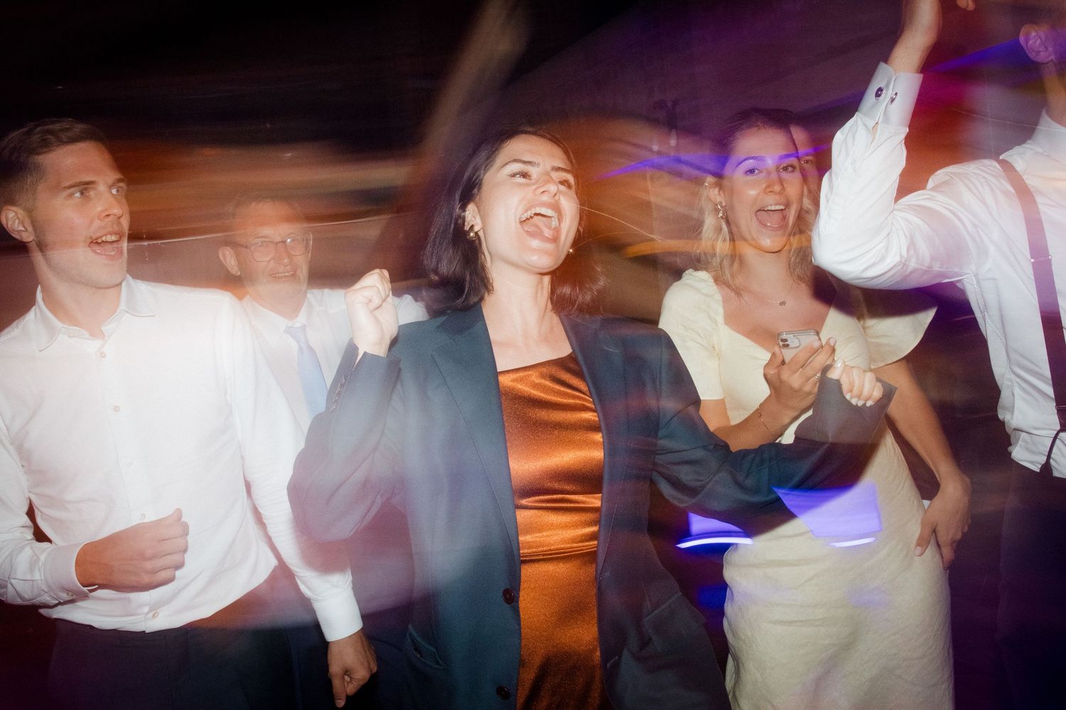 People dancing and celebrating at a lively wedding reception with colorful lighting.