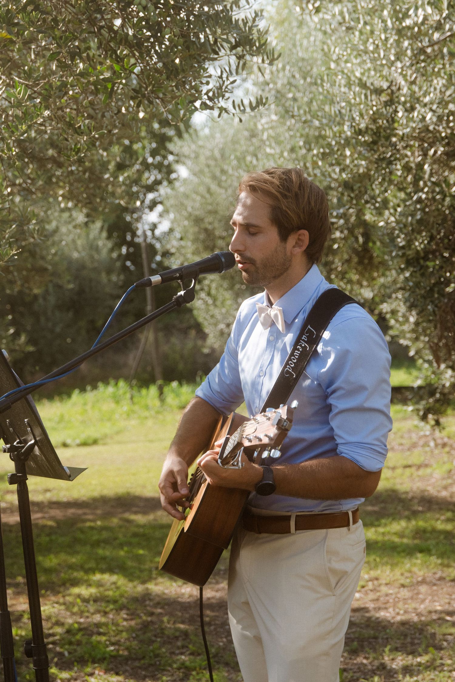 Musician performing with acoustic guitar at outdoor wedding among olive trees.