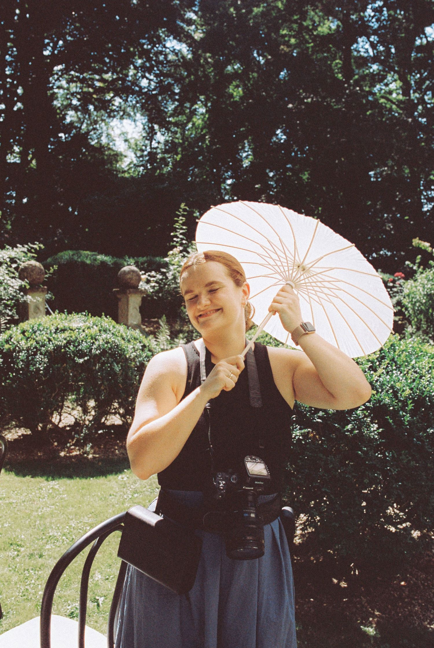 A smiling woman holds a white paper parasol umbrella in a lush garden, wearing a camera and backpack.