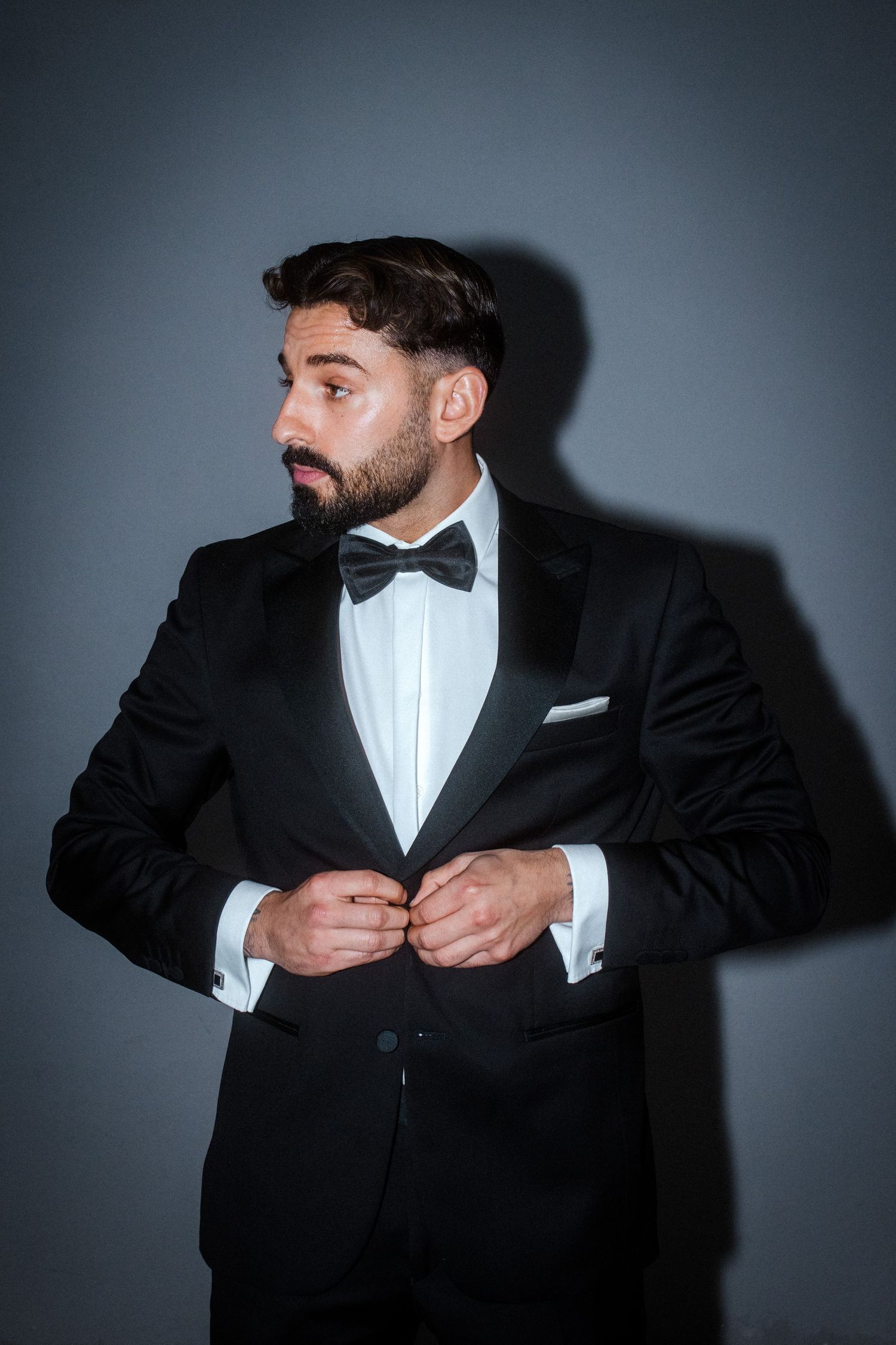 Close-up of groom adjusting his bowtie and tuxedo against grey backdrop.