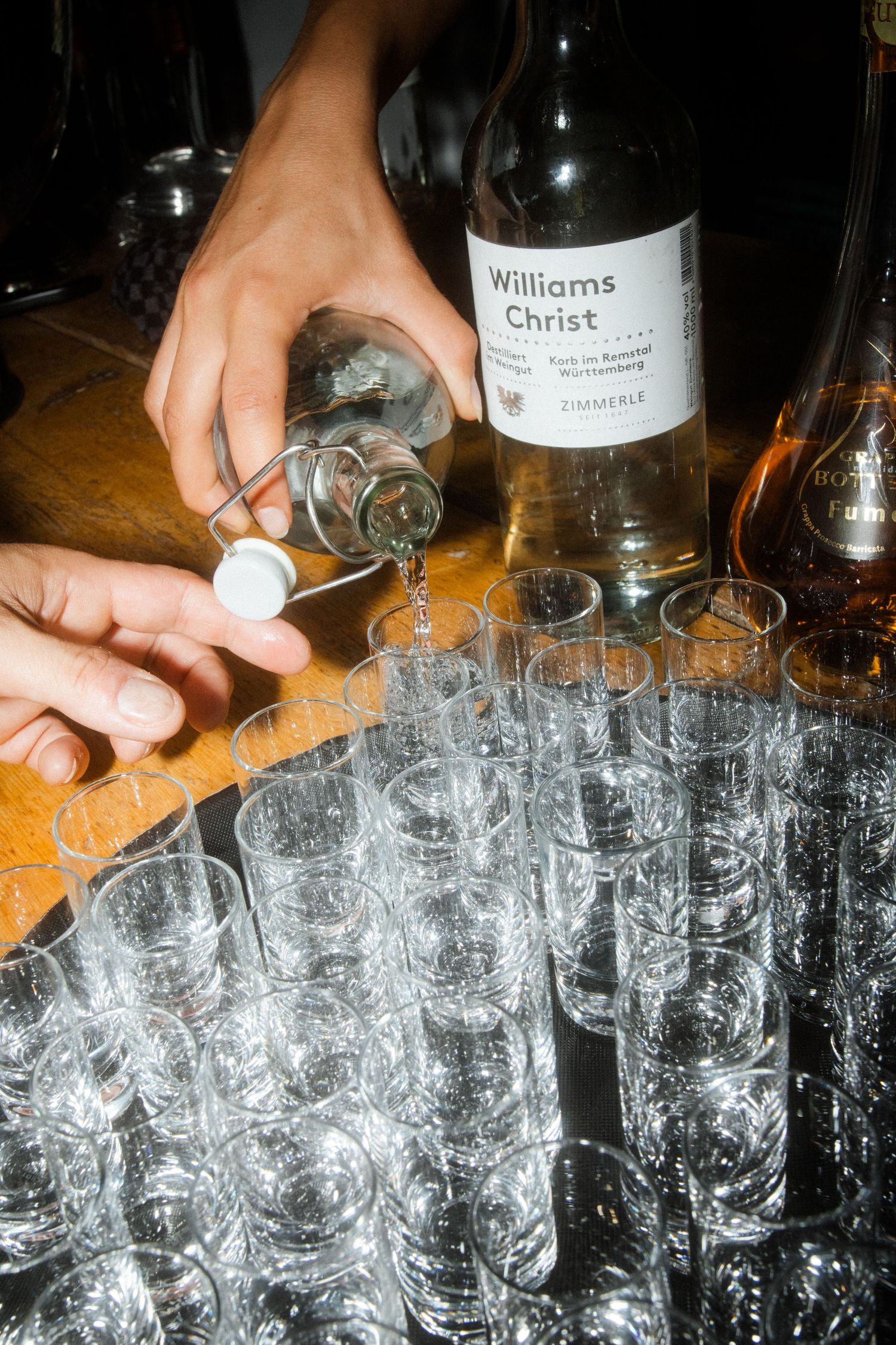 Pouring drinks into multiple shot glasses with Williams Christ bottle on wooden surface.