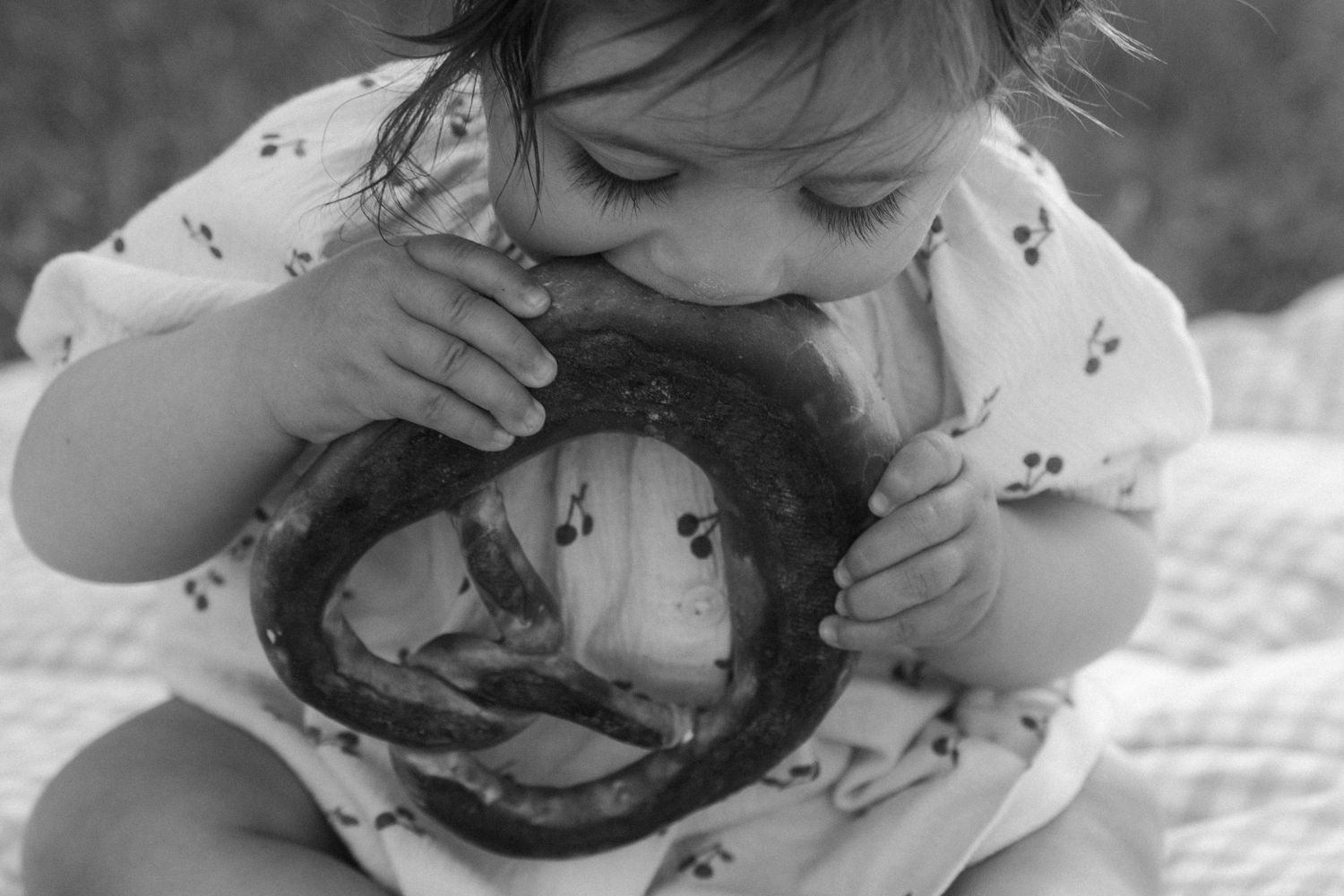 Black and white photo of a baby girl in a floral outfit joyfully eating a large soft pretzel.