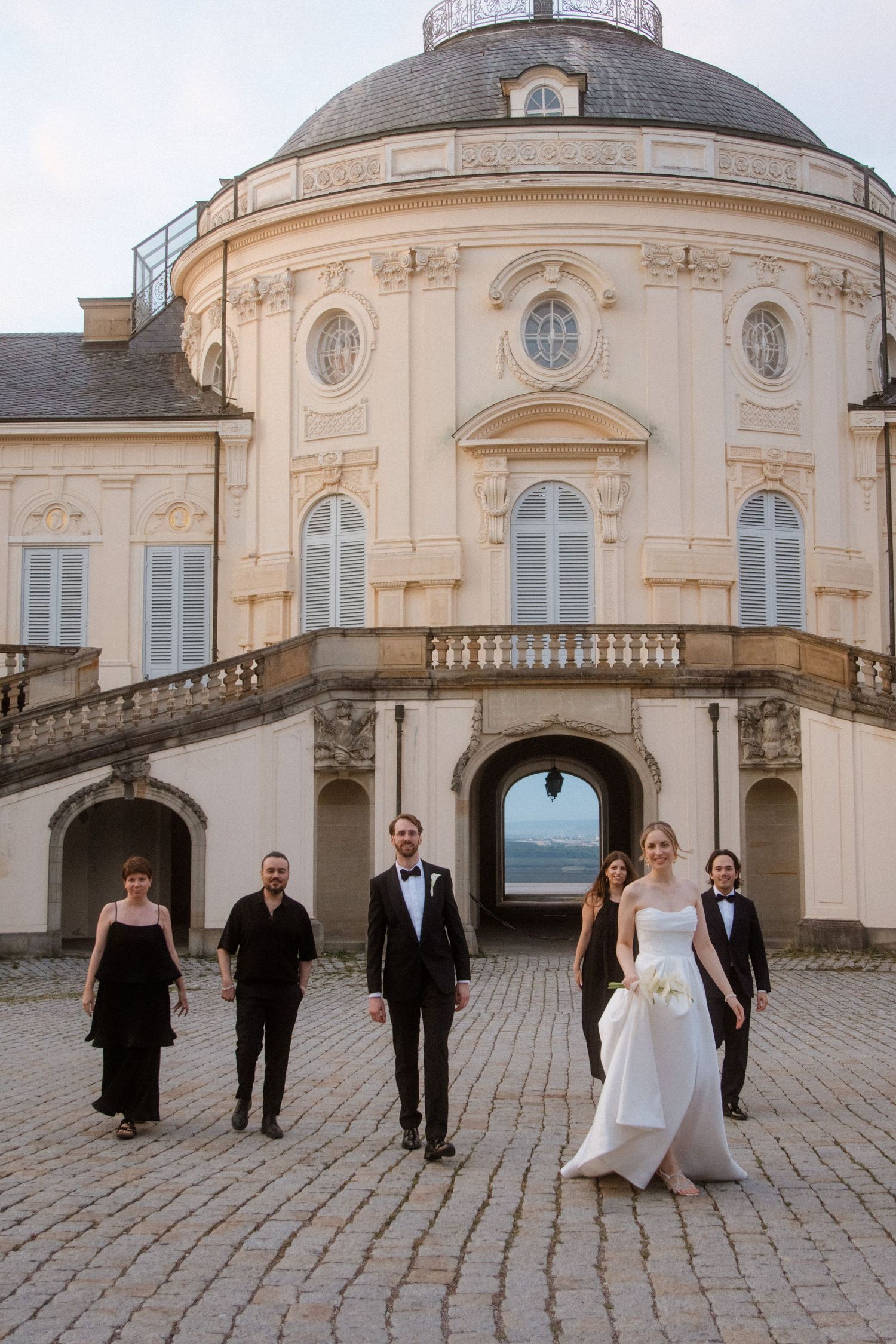 Wedding group poses in front of baroque architecture with curved staircases.