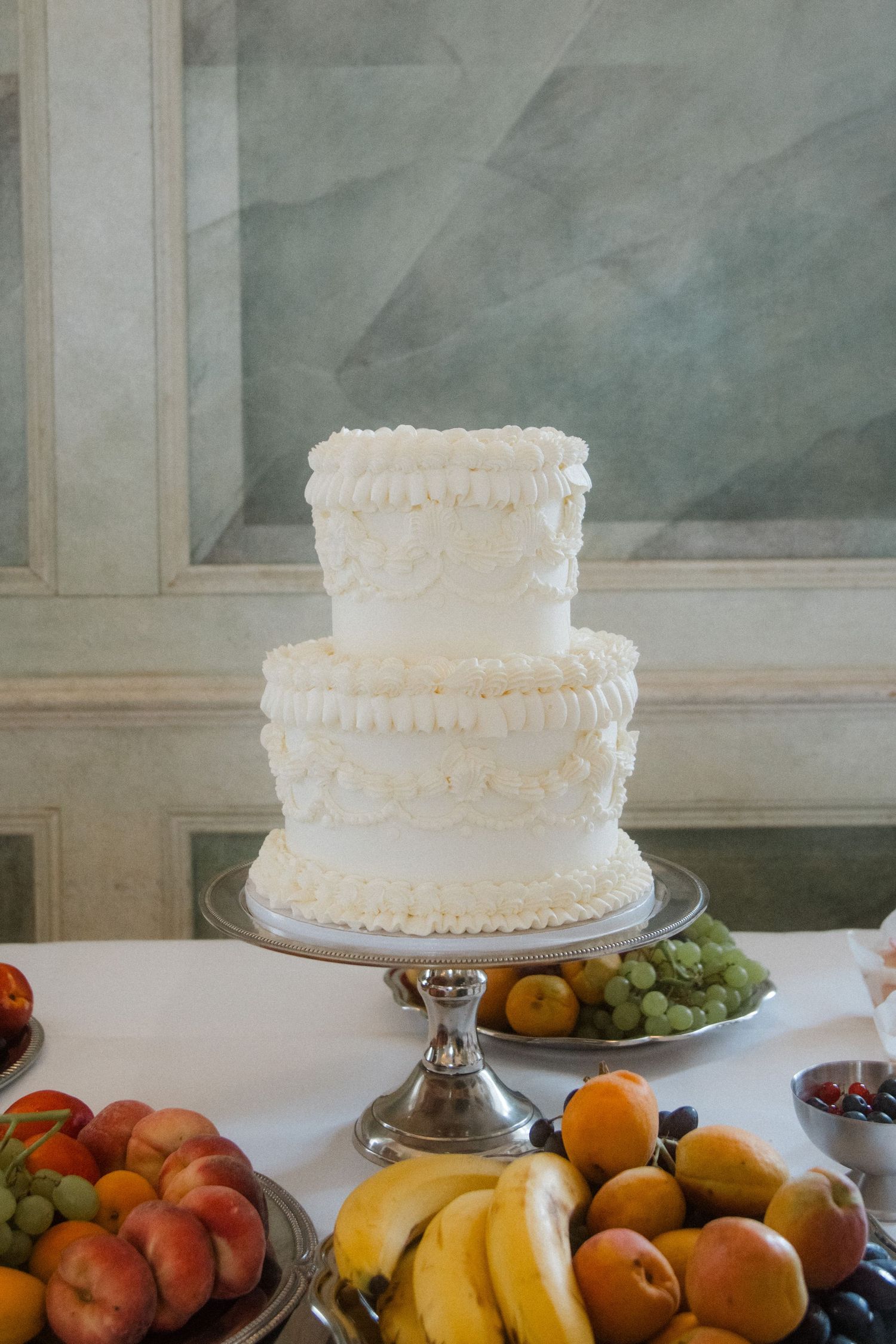Elegant white tiered wedding cake displayed with fresh fruit on a dessert table.
