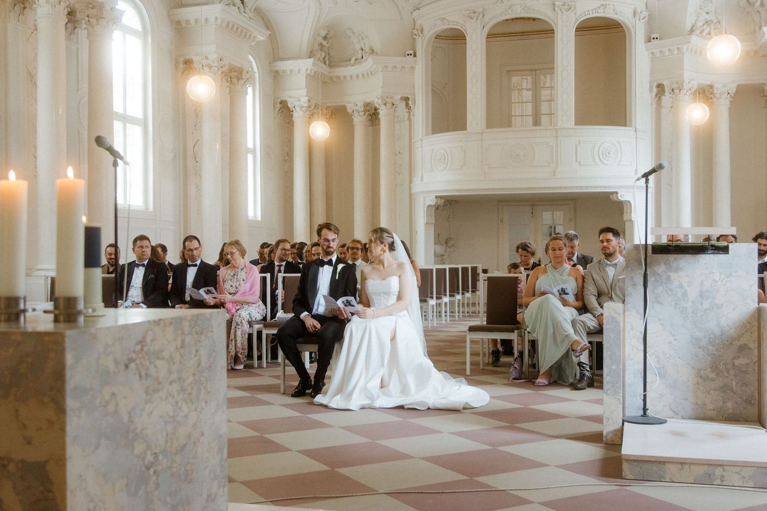 Wedding ceremony in a beautiful baroque-style hall with checkered floors and white columns.