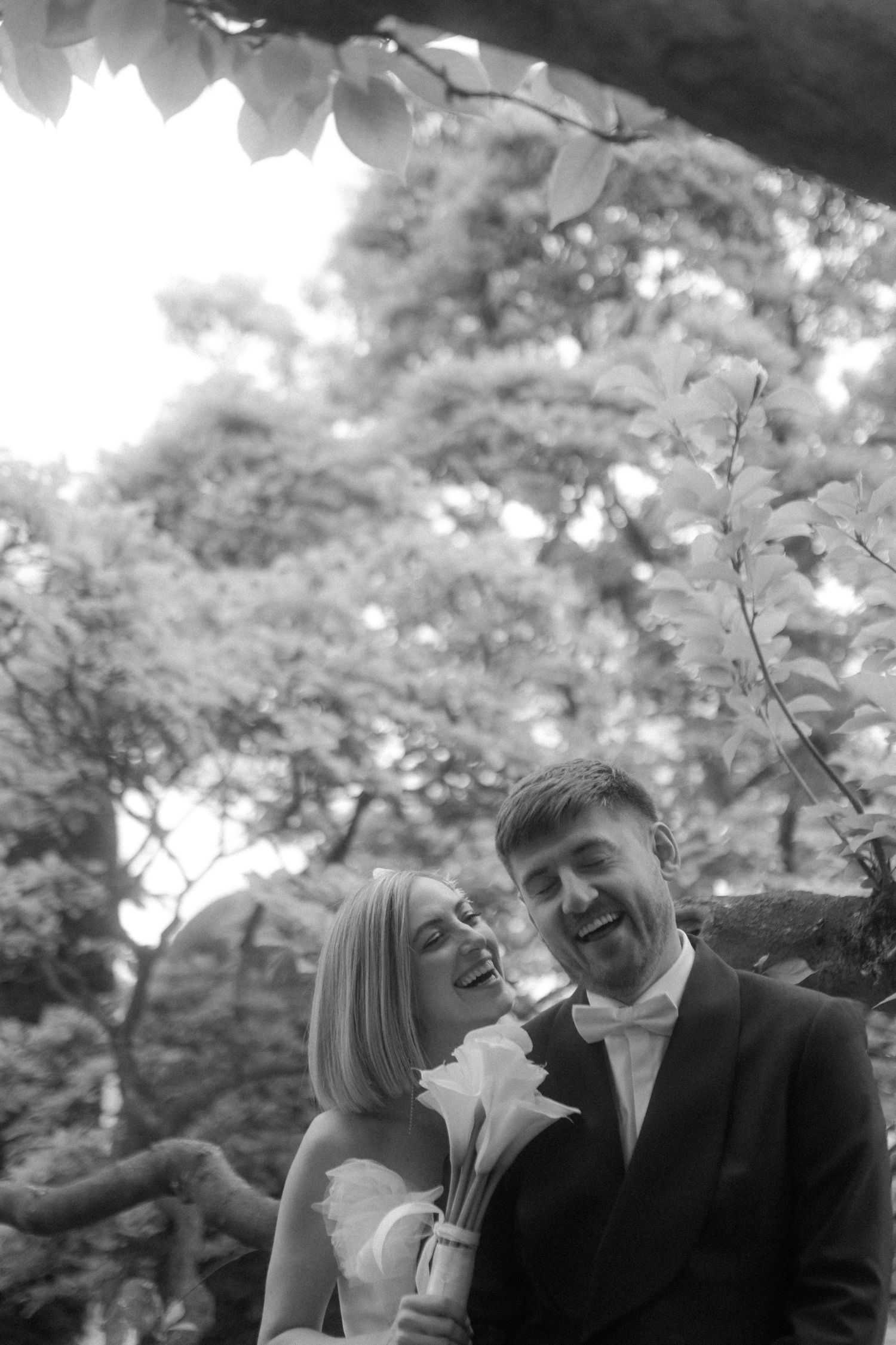 Black and white portrait of a joyful couple sharing a candid moment under garden trees.