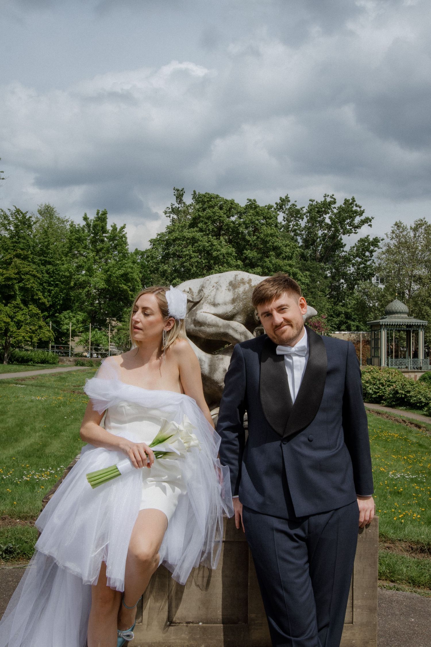 Wedding couple posing by stone statue in garden, bride in modern short dress holding calla lilies.