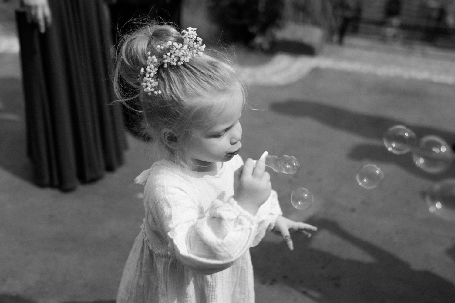 A black and white photo of a young child in a white dress blowing bubbles outdoors.
