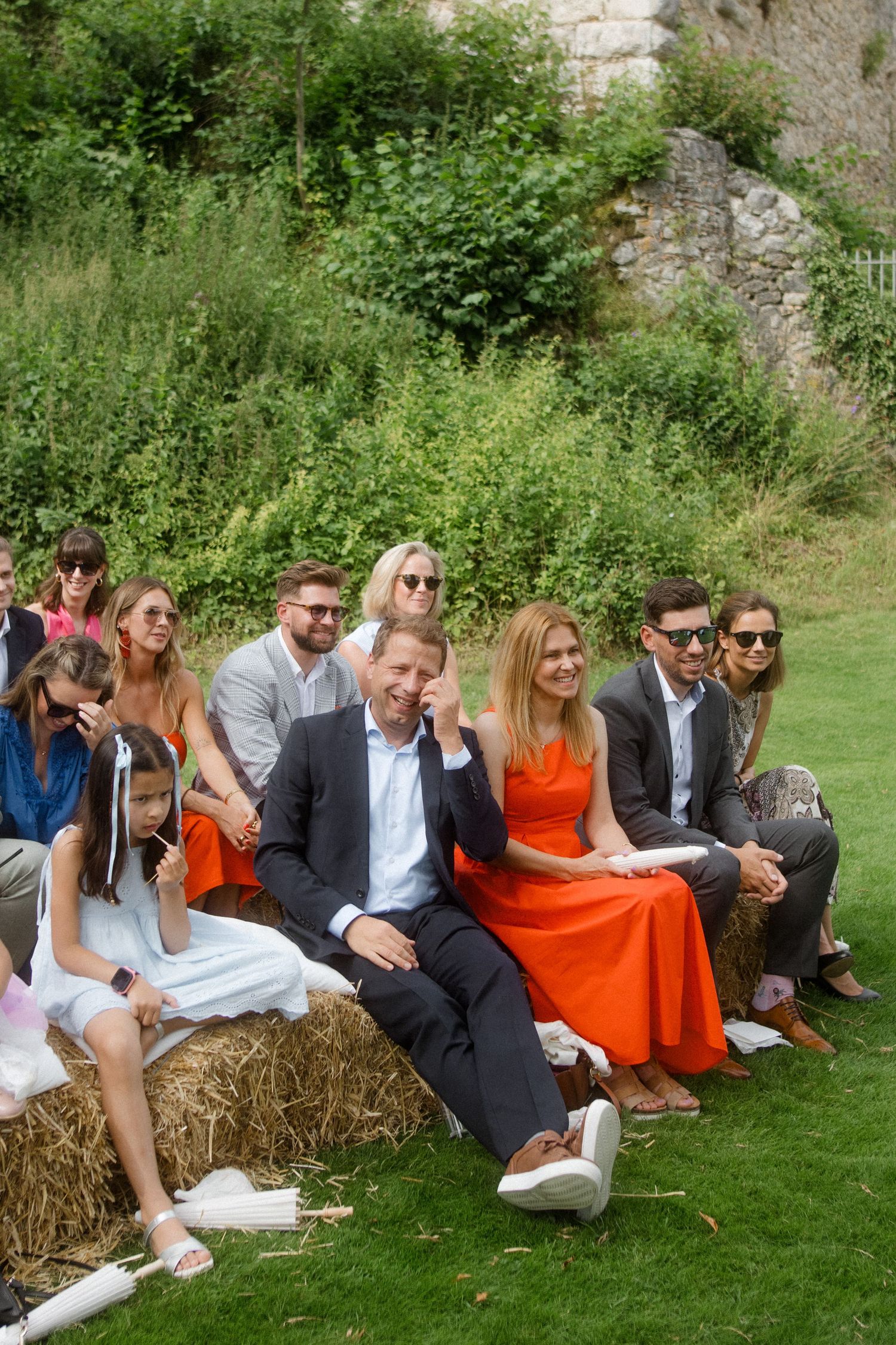 Wedding guests in colorful attire seated on hay bales during outdoor ceremony.