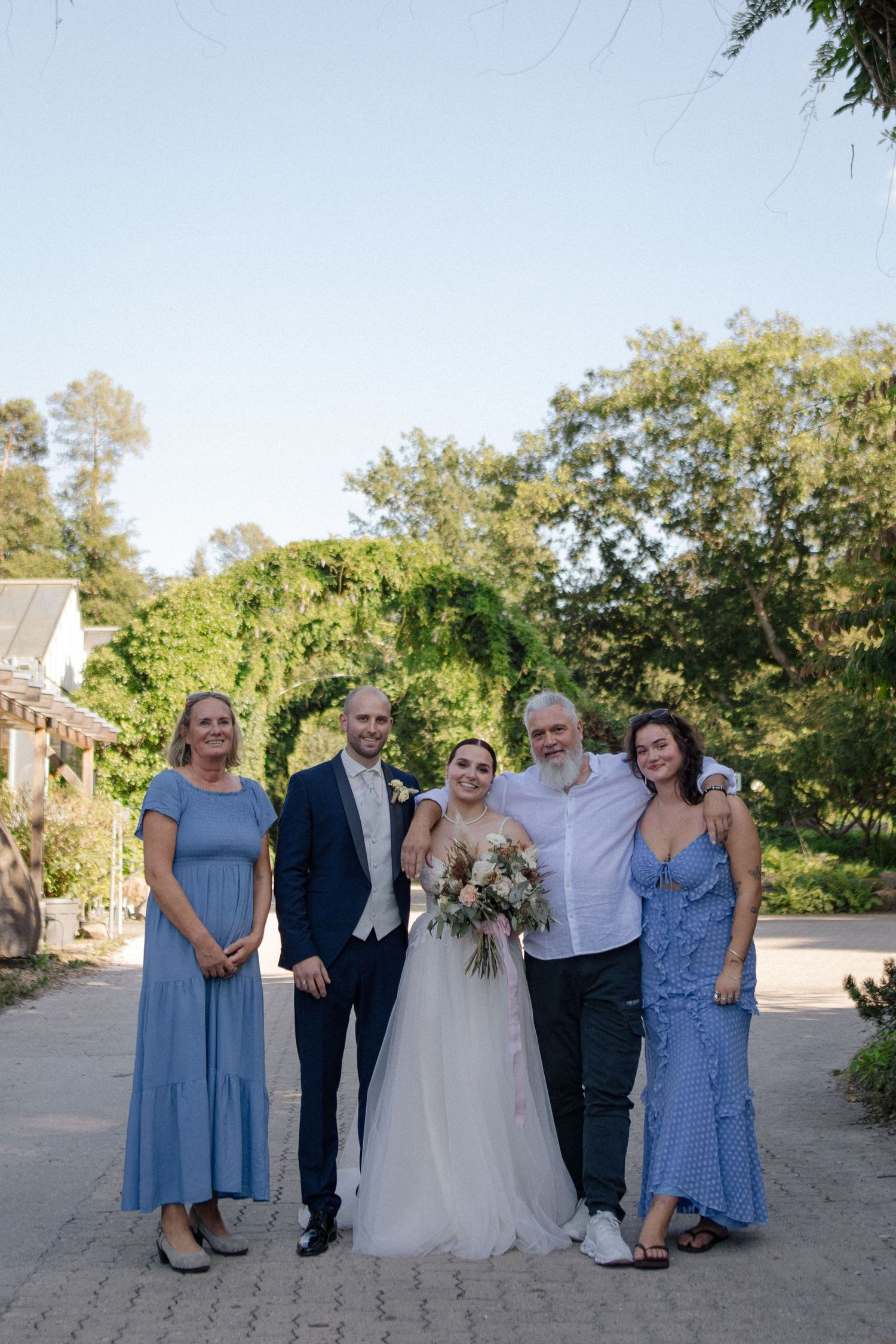 A wedding group photo with five people in formal attire standing on a brick paved outdoor path.