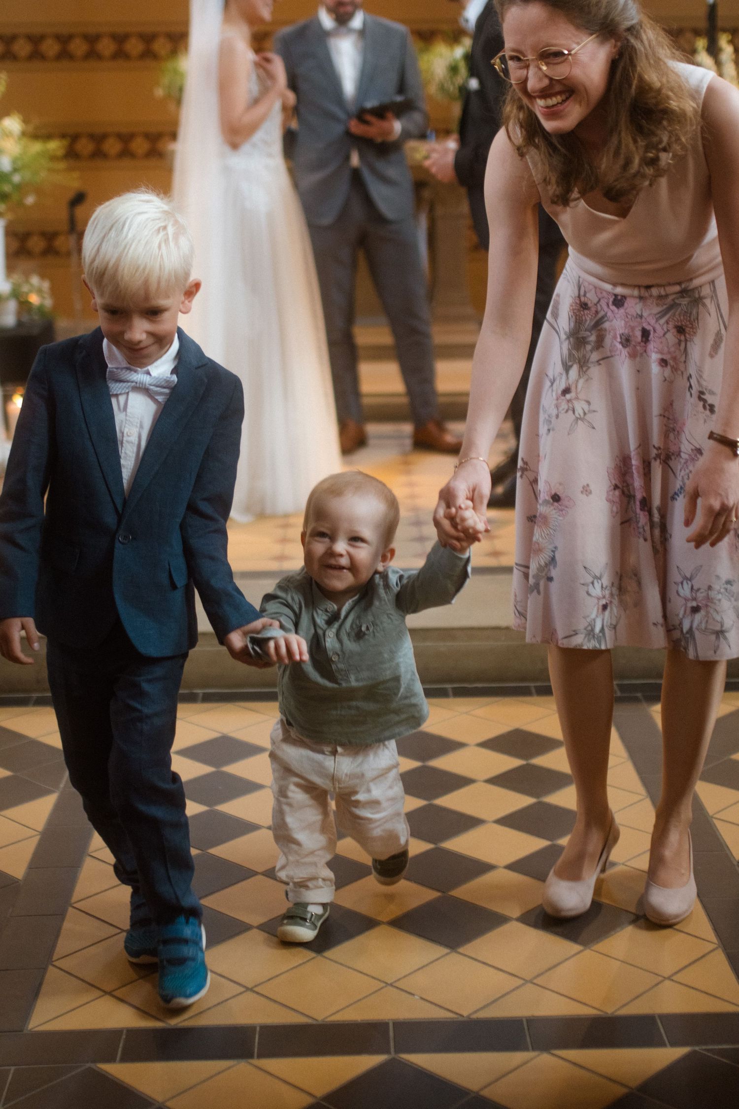 Young children walking with wedding guest on checkered church floor during ceremony.