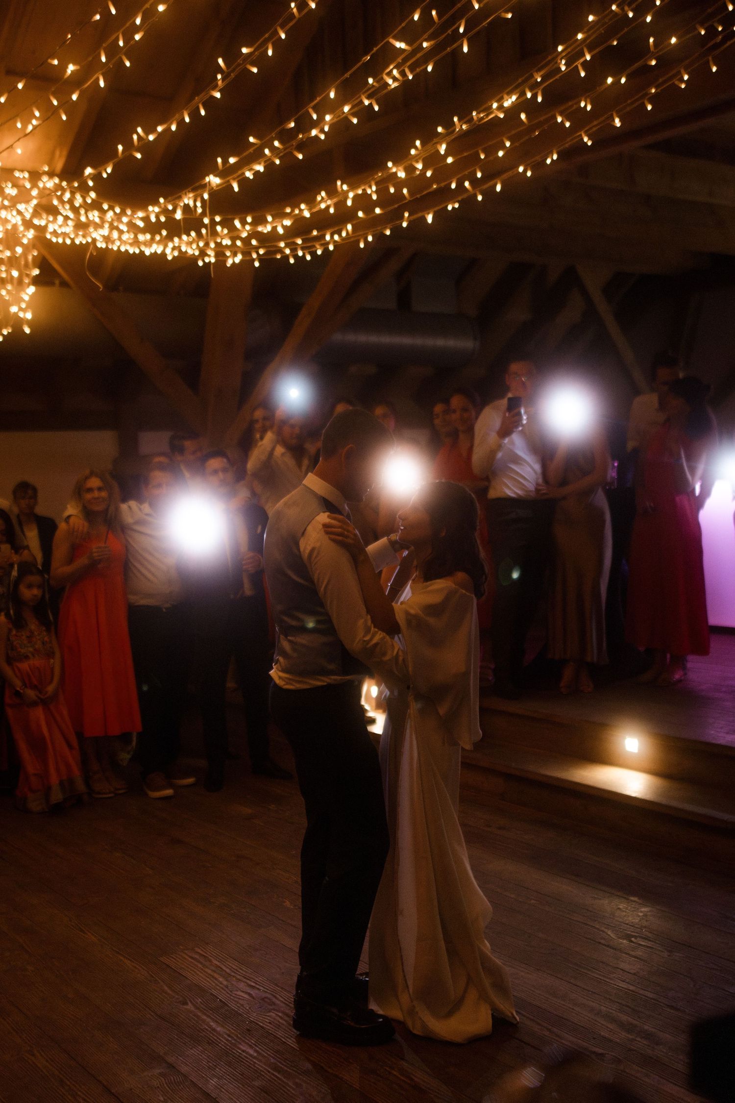 Dancing couple surrounded by guests holding phone lights under warm string light decor.