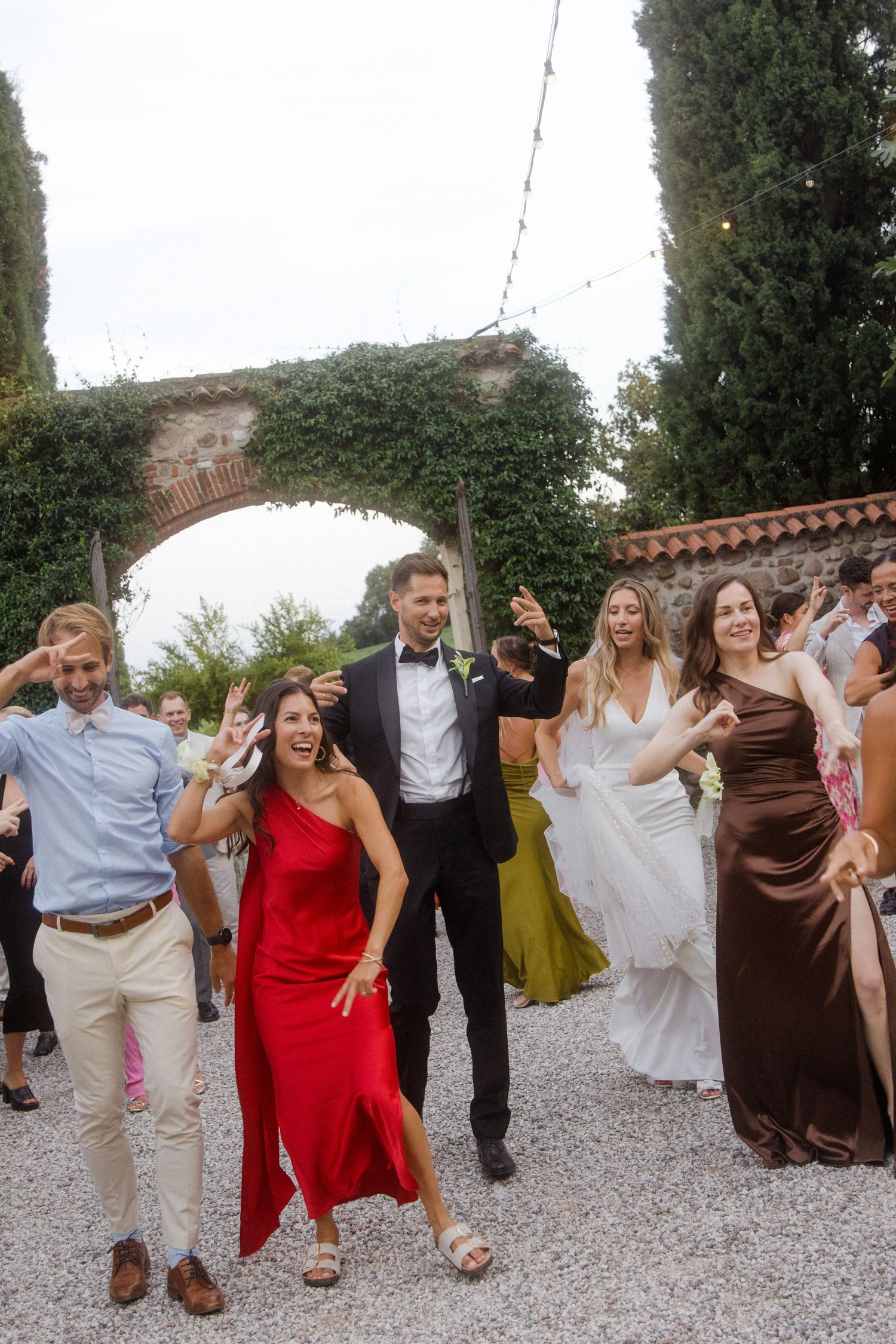 Wedding party dances in coordinated moves on gravel courtyard during outdoor reception.