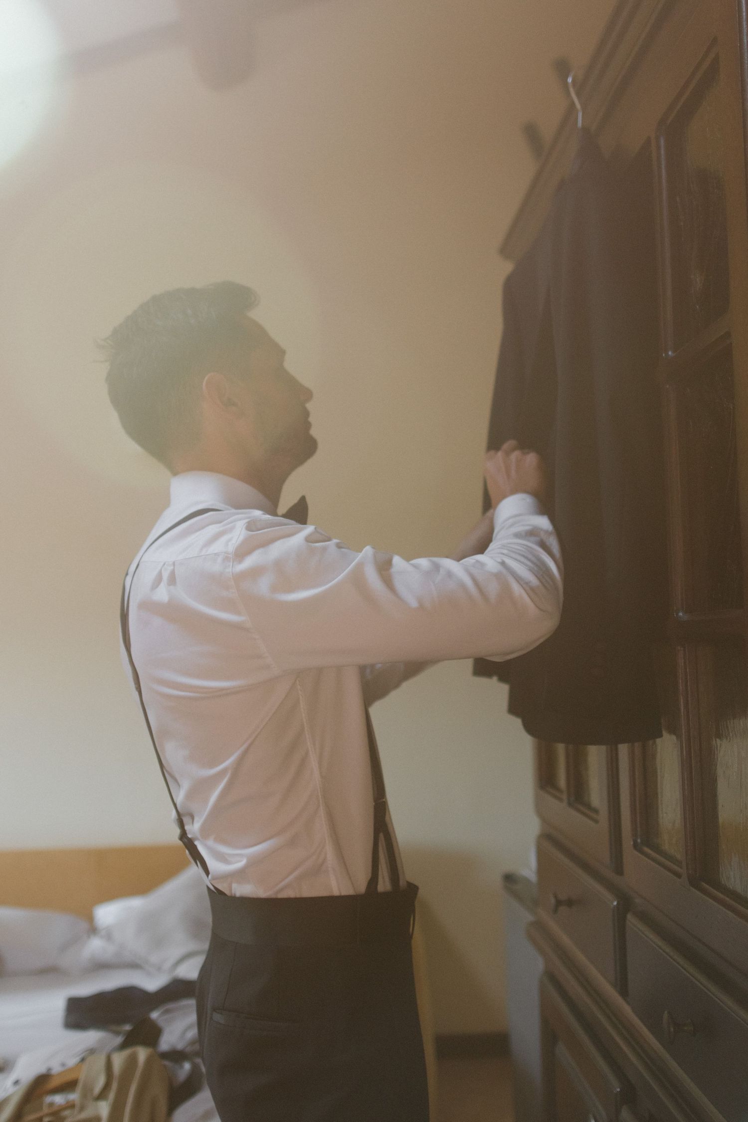 Someone in white shirt and suspenders reaches toward a cabinet in a sunlit room.