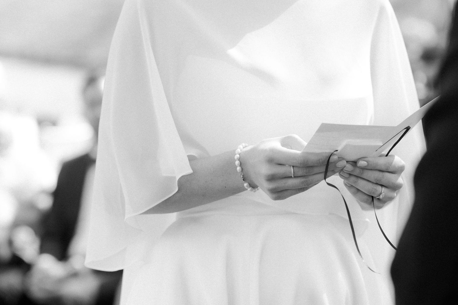 Bride in white dress reading wedding vows from a card, wearing a pearl bracelet, black and white photo.