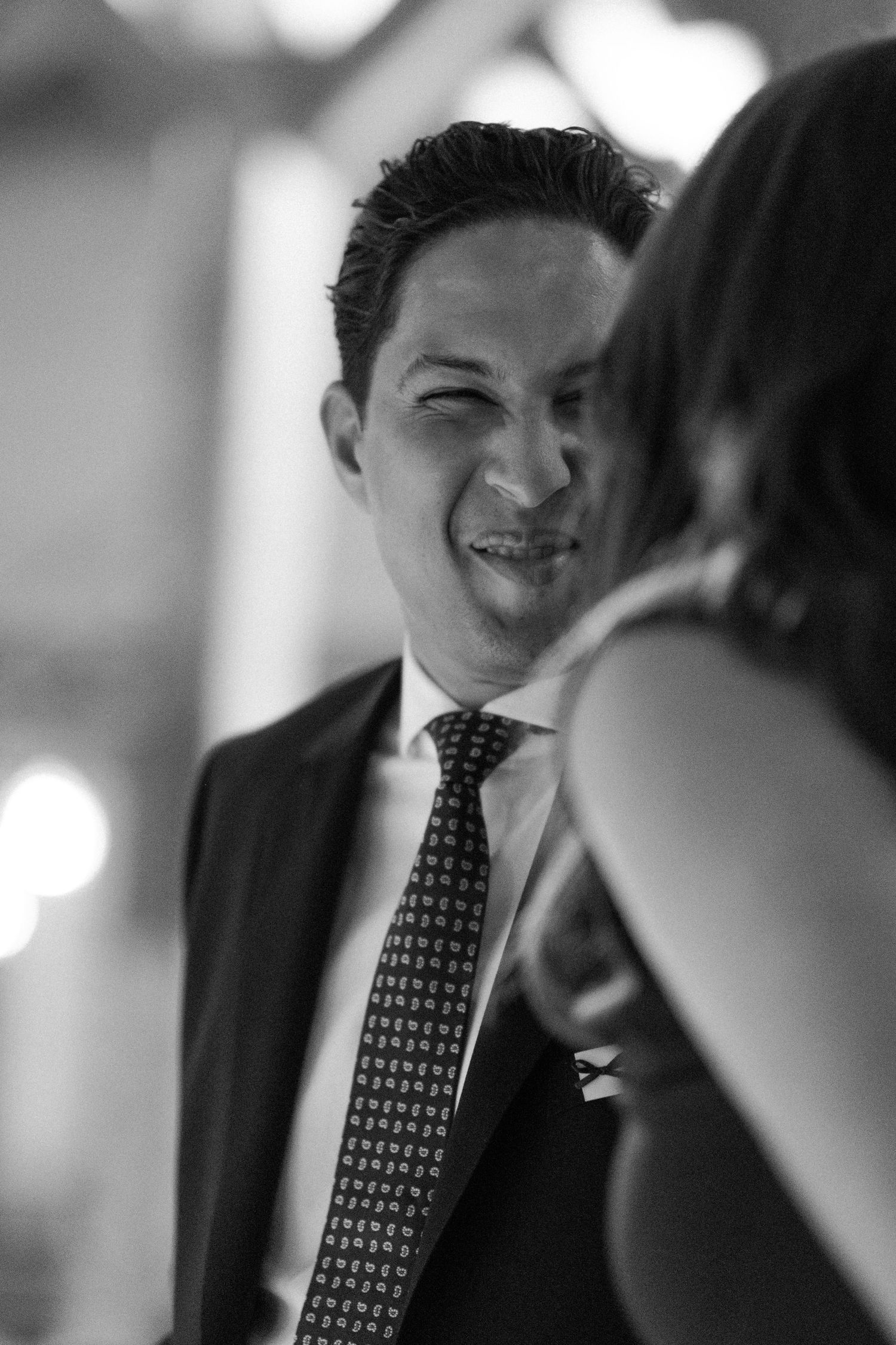 A man in a suit and patterned tie smiles warmly at a woman during a wedding reception in black and white.