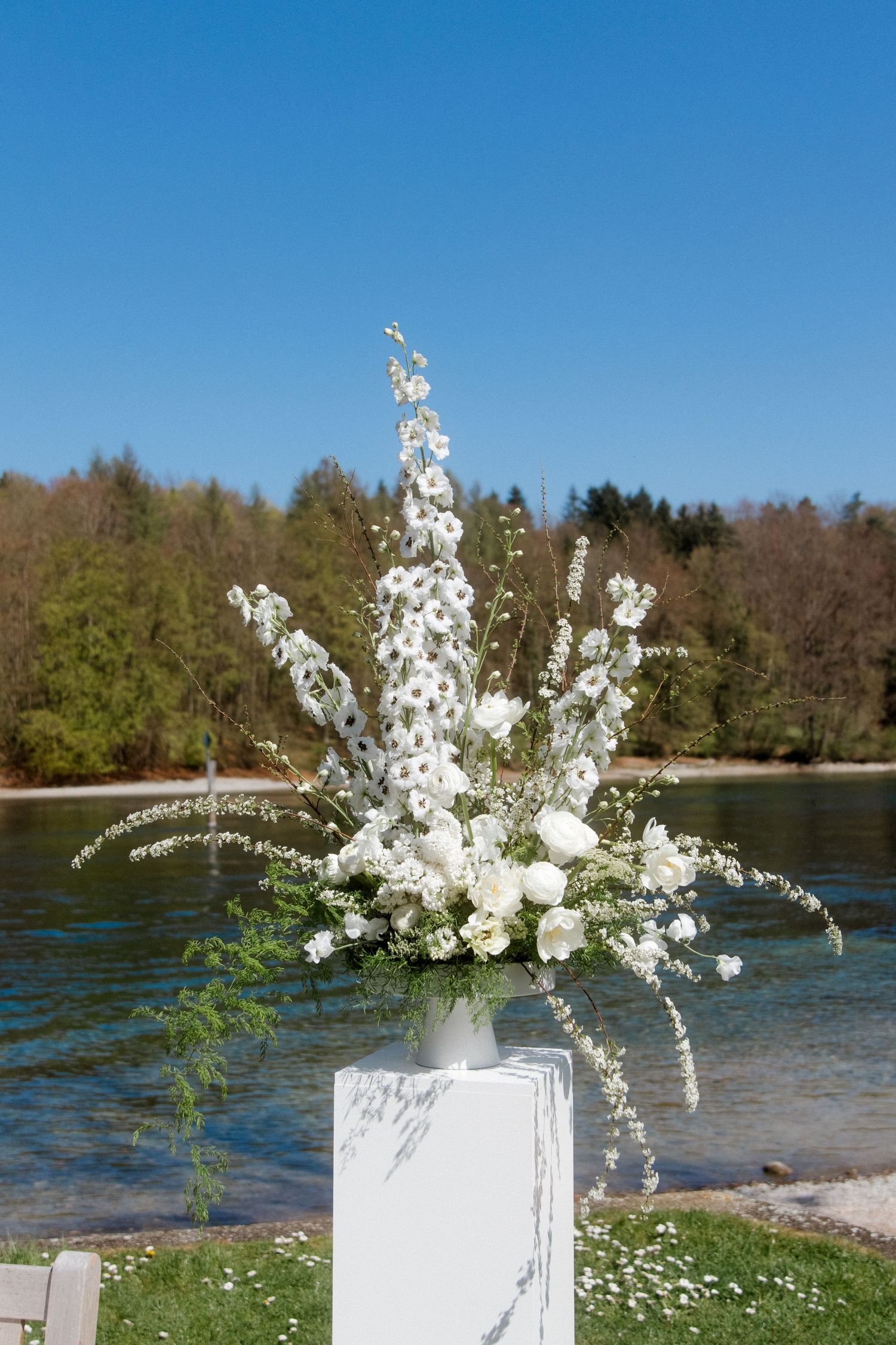 White floral arrangement with delphiniums and roses on a pedestal by a scenic lakeside on a sunny day.