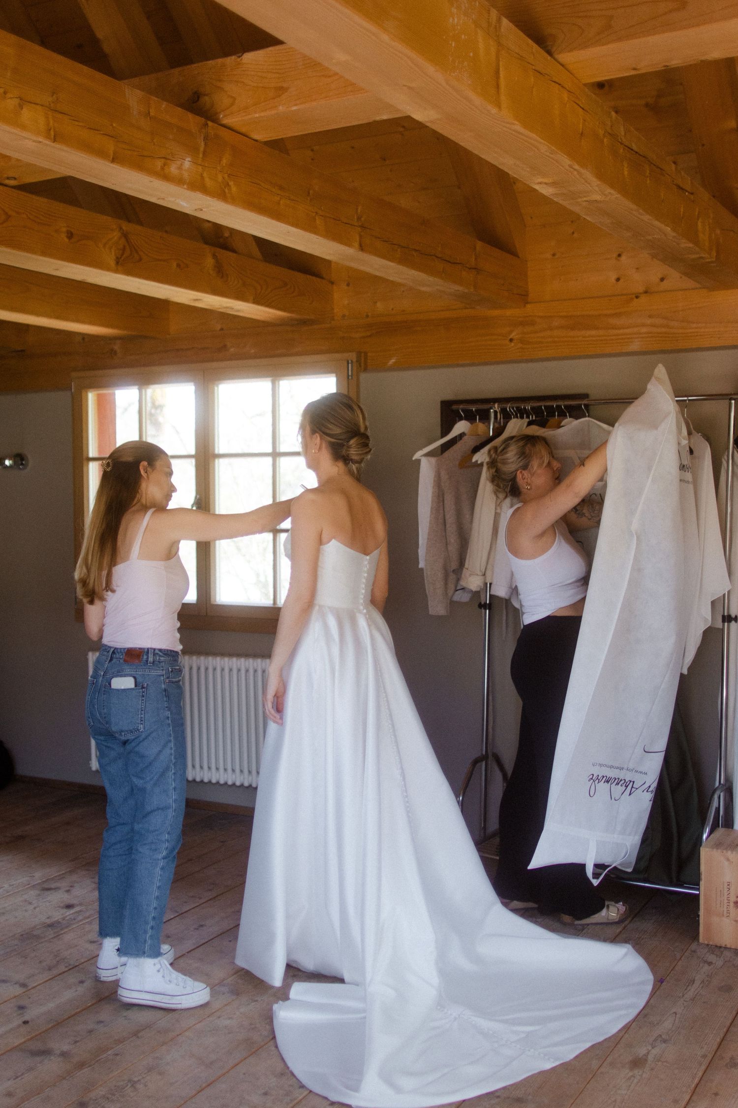 Two women helping a bride into her white wedding gown in a rustic wooden room.