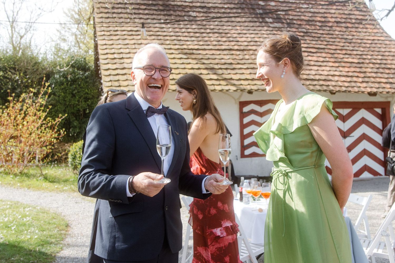 Man in tuxedo and woman in green dress laughing outdoors at elegant garden party with champagne glasses.